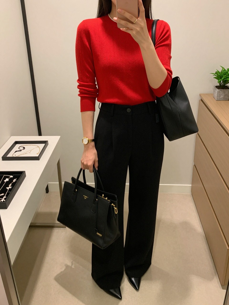 mirror selfie of a Latina woman wearing a red fitted knit top, black high-waisted wide-leg trousers, black pointed pumps, gold watch, and a black structured tote bag, holding a phone in front of a mirror, cropped at NECK so the face does not show, relaxed casual selfie pose, in a walk-in closet mirror with soft indoor lighting, including a jewelry tray on the vanity and a small plant on a dresser, realistic latest iPhone photo quality.