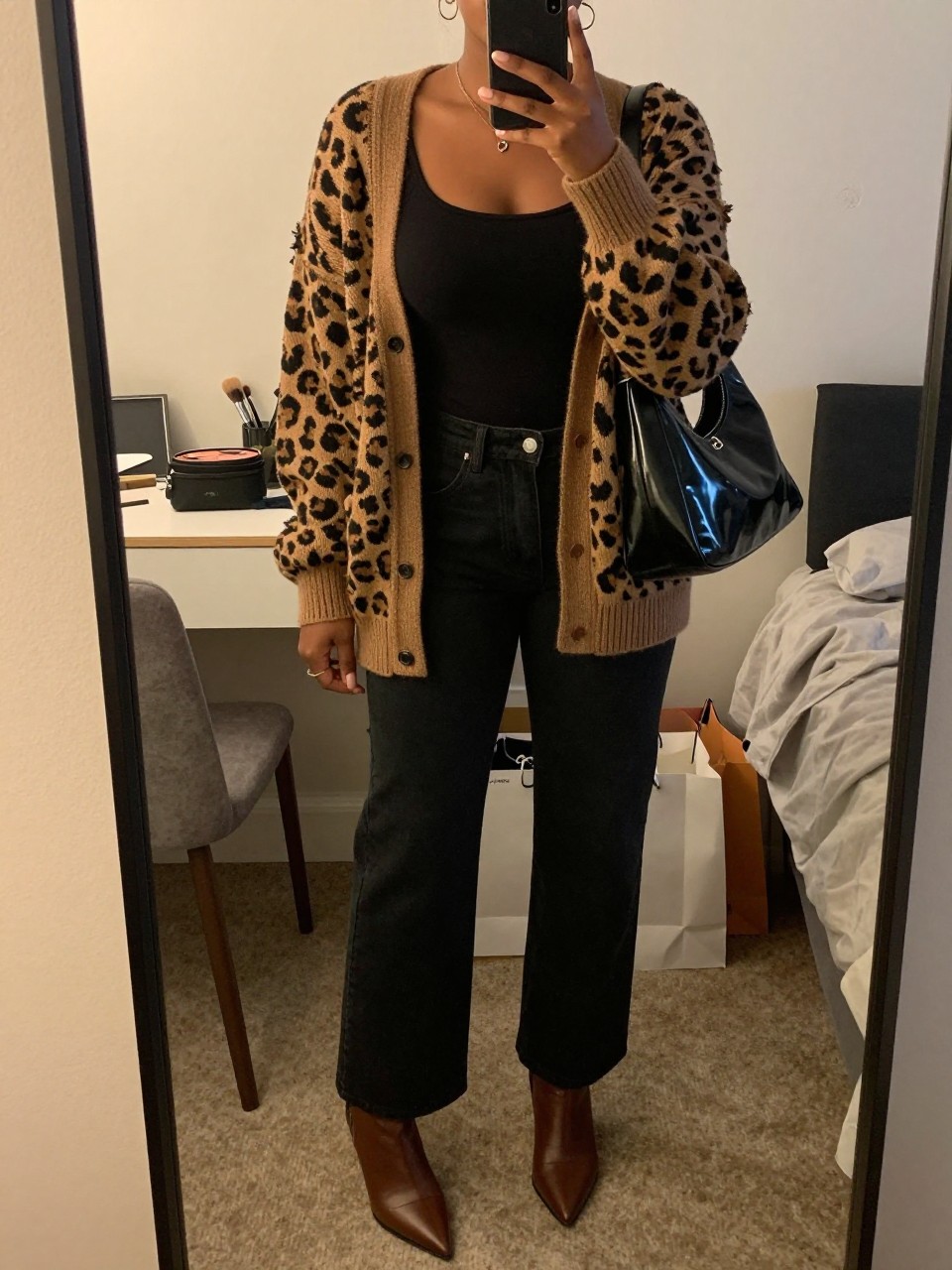 mirror selfie of a Black woman wearing the exact outfit described above, clearly showing a brown leopard knit cardigan, fitted black tank, black wide-leg jeans, brown pointed ankle boots, and a glossy black shoulder bag, holding a phone in front of a mirror, cropped at NECK so the face does not show, with a relaxed casual selfie pose, in a cozy bedroom mirror setup, evening golden hour, with a makeup brush or cosmetic bag on the counter and shopping bags beside a chair, latest iPhone photo quality.