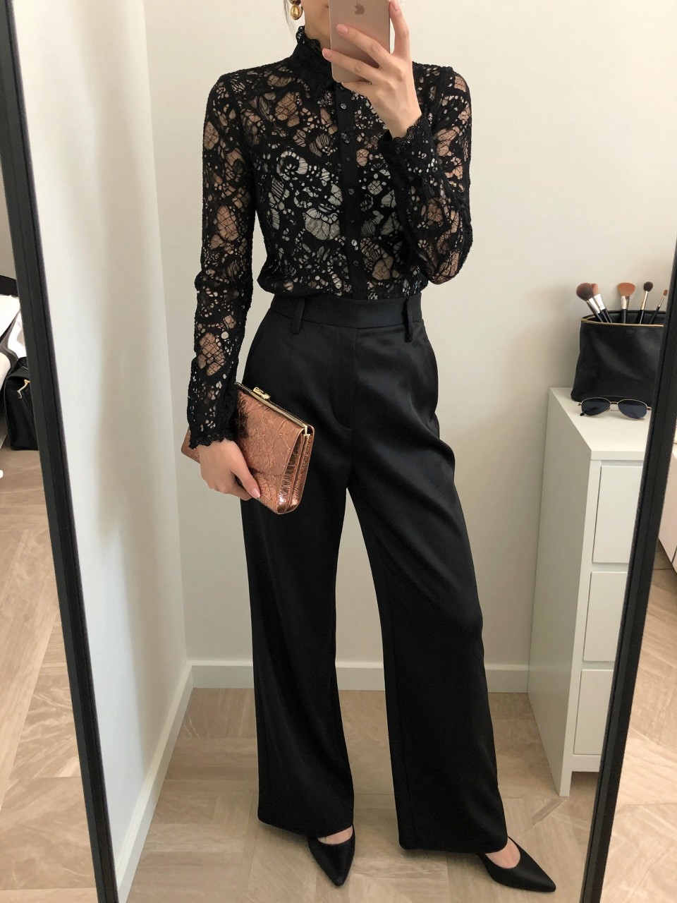 mirror selfie of a woman wearing the exact outfit described above, clearly showing a black lined lace blouse with long sleeves, black satin wide-leg trousers, black pointed pumps, a metallic bronze clutch, and gold drop earrings, holding a phone in front of a mirror, cropped at NECK so the face does not show, with a relaxed casual selfie pose, in a feminine-themed bedroom, soft indoor lighting, with a makeup brush or cosmetic bag on the counter and sunglasses placed on a dresser, latest iPhone photo quality.