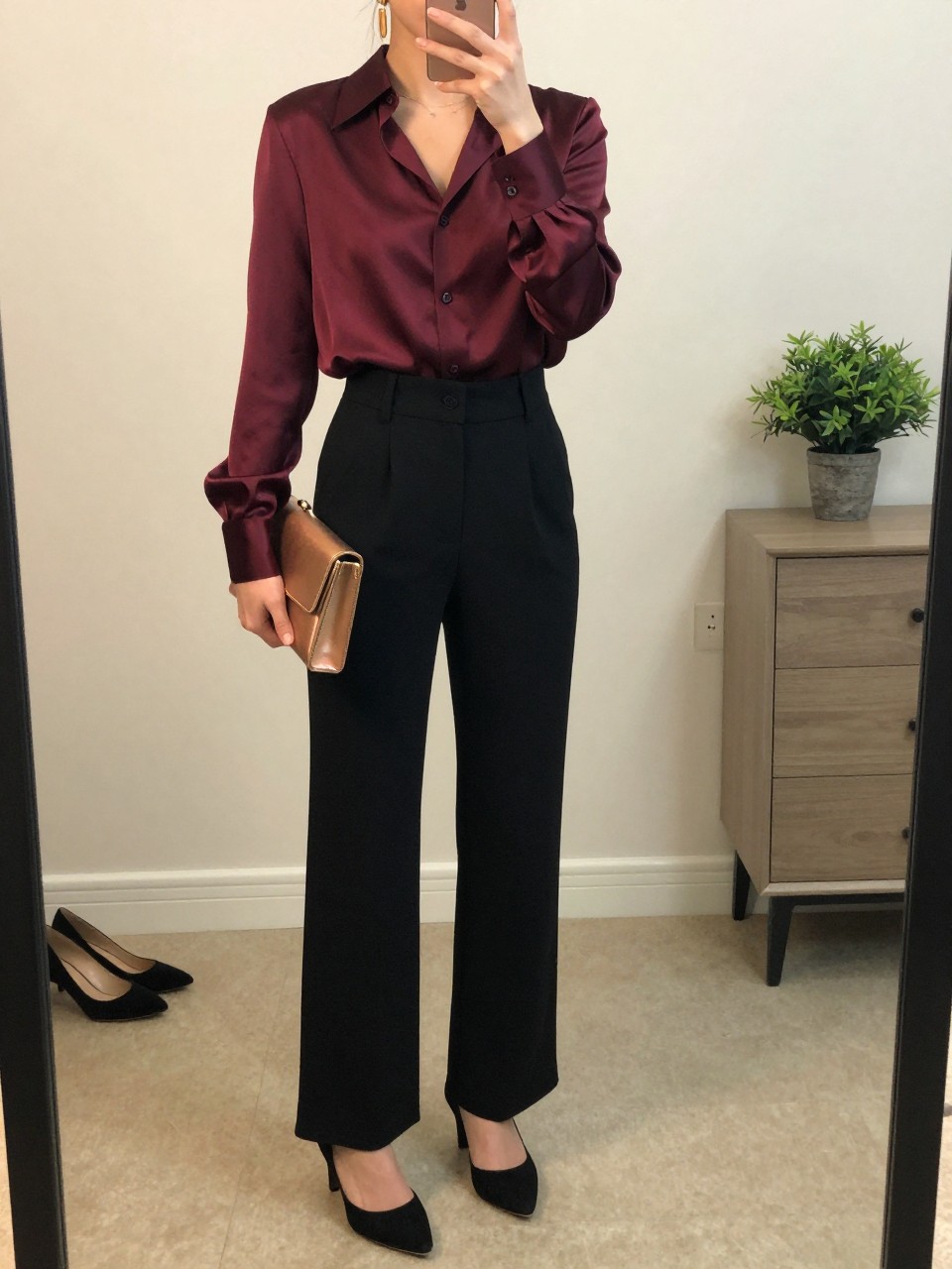 mirror selfie of a woman wearing the exact outfit described above, clearly showing a deep plum satin blouse, black high-waisted wide-leg trousers, black suede pumps, gold drop earrings, and a bronze clutch, holding a phone in front of a mirror, cropped at NECK so the face does not show, with a relaxed casual selfie pose, in a minimal dressing room, soft indoor lighting, with a small plant on a dresser and a pair of heels on the floor nearby, latest iPhone photo quality.