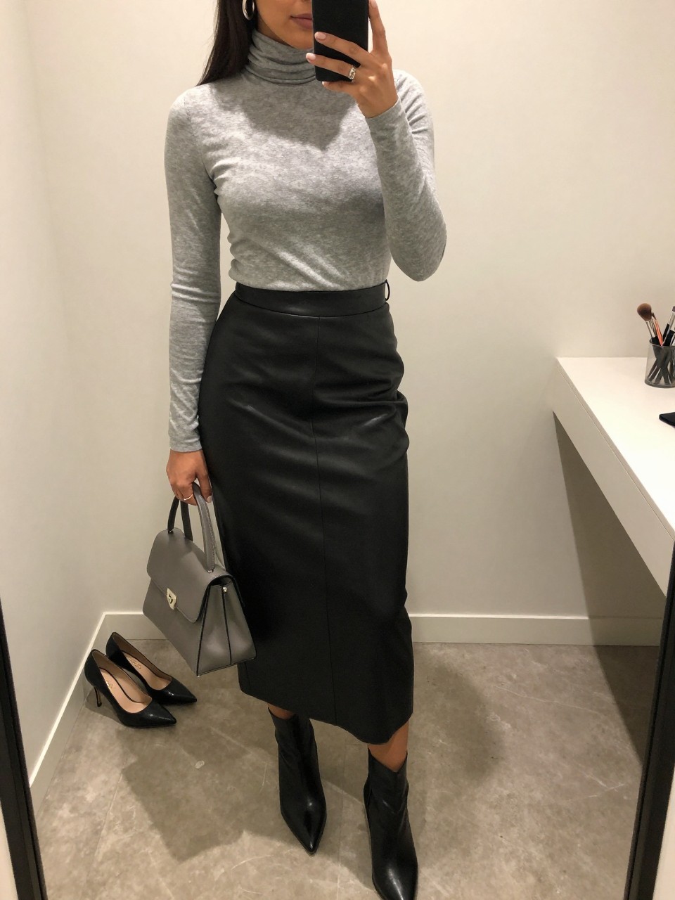 mirror selfie of an olive-toned Latina woman wearing a soft gray fitted turtleneck, black leather midi skirt, black pointed ankle boots, silver hoop earrings, and a gray mini handbag, holding a phone in front of a mirror, cropped at NECK so the face does not show, relaxed casual selfie pose, in a minimal dressing room with soft indoor lighting, including a pair of heels on the floor nearby and a makeup brush on the counter, realistic latest iPhone photo quality.