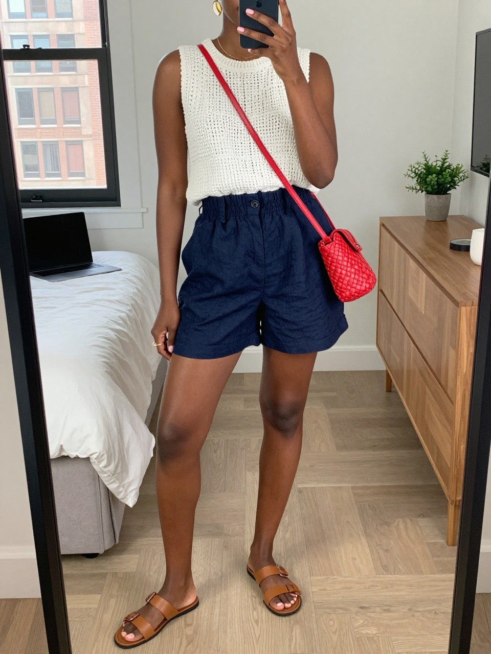 mirror selfie of a Black woman wearing a white sleeveless crochet top, navy high-waisted linen shorts, tan leather slide sandals, red woven crossbody bag, and gold shell earrings, clearly showing the same clothing items, colors, fabrics, and accessories listed in the Outfit Pieces section, holding a phone in front of a mirror, cropped at NECK so the face does not show, with a relaxed casual selfie pose, in a stylish city apartment, natural window light, including a laptop on the bed and a small plant on a dresser, latest iPhone photo quality.