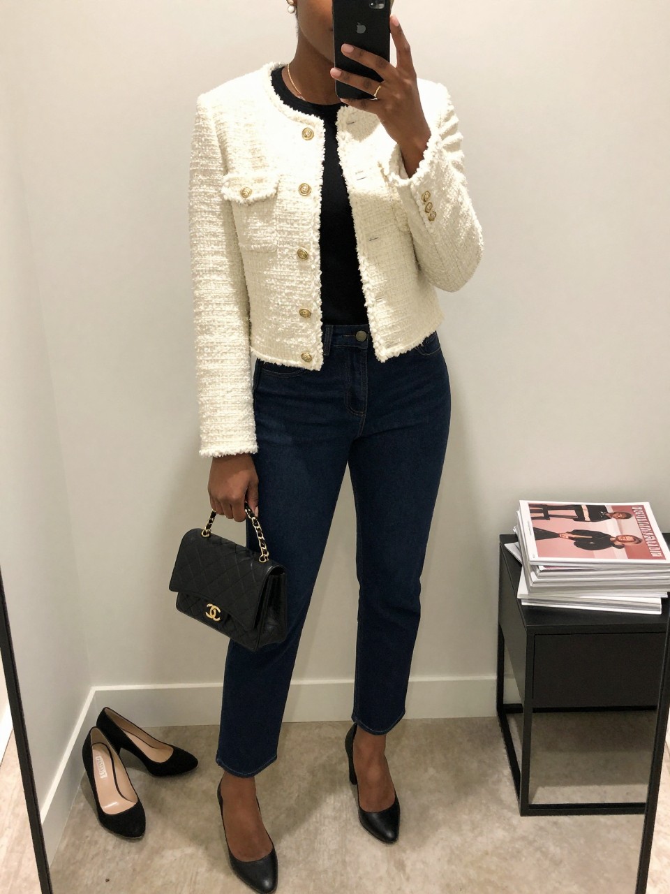 mirror selfie of a Black woman wearing the exact outfit described above, clearly showing a cream cropped tweed jacket layered over a black shell top, dark indigo trouser jeans, black block-heel pumps, a black quilted mini bag, and pearl stud earrings, holding a phone in front of a mirror, cropped at NECK so the face does not show, with a relaxed casual selfie pose, in a minimal dressing room, soft indoor lighting, with a pair of heels on the floor nearby and a stack of fashion magazines on a side table, latest iPhone photo quality.