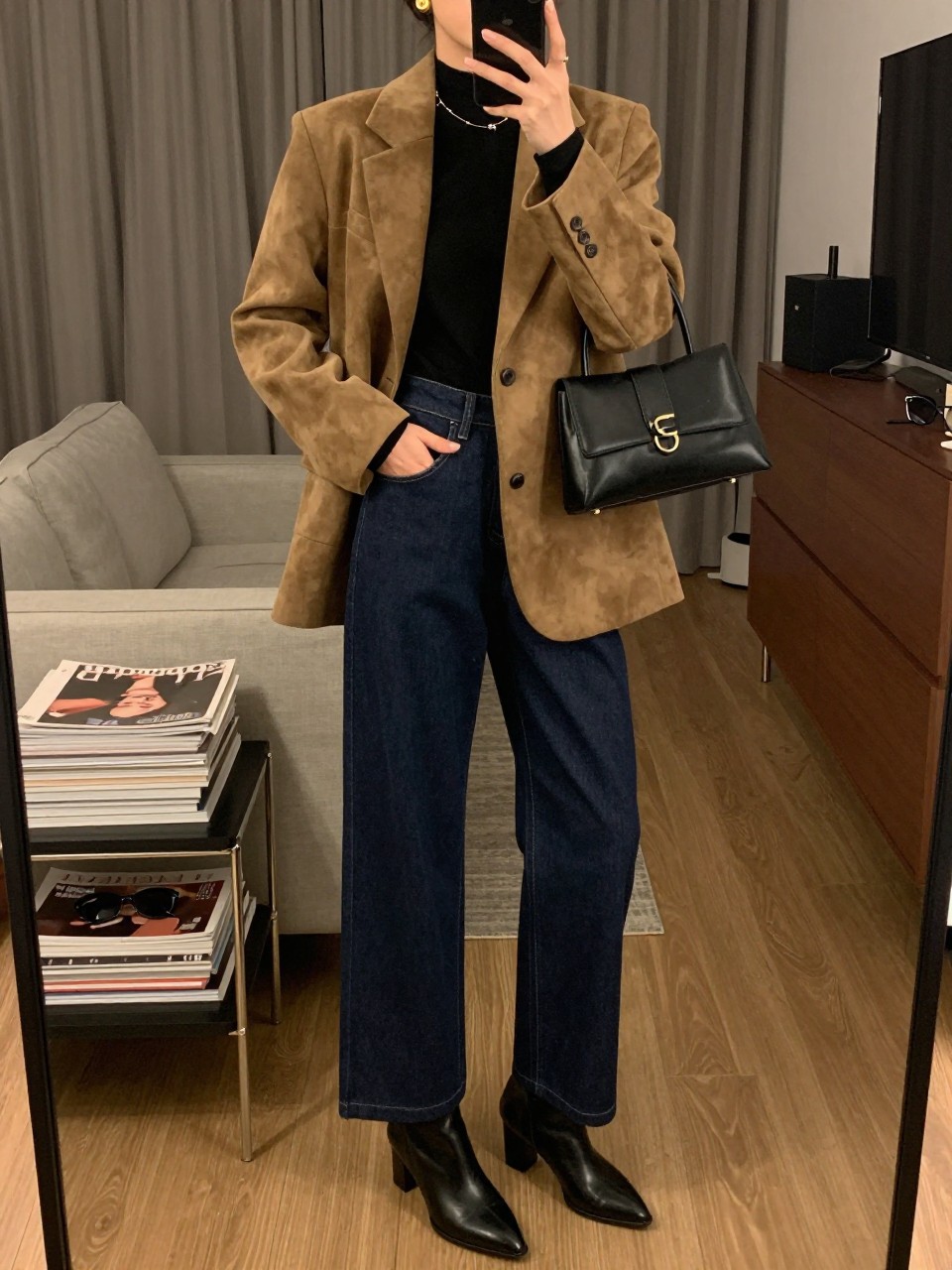 mirror selfie of a woman wearing the exact outfit described above, clearly showing dark blue wide-leg denim trousers, a camel suede blazer layered over a fitted black mock-neck top, black heeled ankle boots, a structured black handbag, and gold drop earrings, holding a phone in front of a mirror, cropped at NECK so the face does not show, with a relaxed casual selfie pose, in a stylish city apartment, warm moody lighting, with a stack of fashion magazines on a side table and sunglasses placed on a dresser, latest iPhone photo quality.