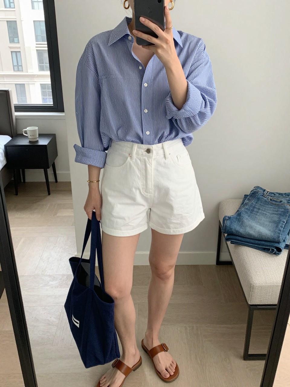 mirror selfie of a woman wearing a blue and white striped oversized button-down shirt tucked loosely into white high-rise denim shorts, brown leather flat sandals, navy canvas tote bag, and chunky gold hoop earrings, clearly showing the same clothing items, colors, fabrics, and accessories listed in the Outfit Pieces section, holding a phone in front of a mirror, cropped at NECK so the face does not show, with a relaxed casual selfie pose, in a stylish city apartment, natural window light, including a coffee mug on a bedside table and folded denim jeans on a bench, latest iPhone photo quality.