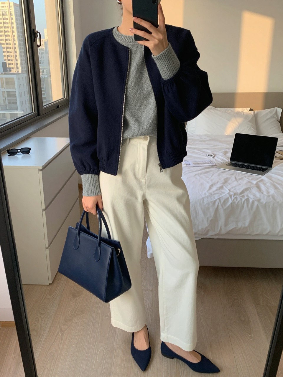 mirror selfie of a woman wearing the exact outfit described above, clearly showing a navy funnel-neck jacket layered over a soft gray fine-knit sweater, ivory wide-leg trousers, navy pointed flats, a structured navy handbag, and silver stud earrings, holding a phone in front of a mirror, cropped at NECK so the face does not show, with a relaxed casual selfie pose, in a stylish city apartment, evening golden hour, with a laptop or tablet on the bed and sunglasses placed on a dresser, latest iPhone photo quality.