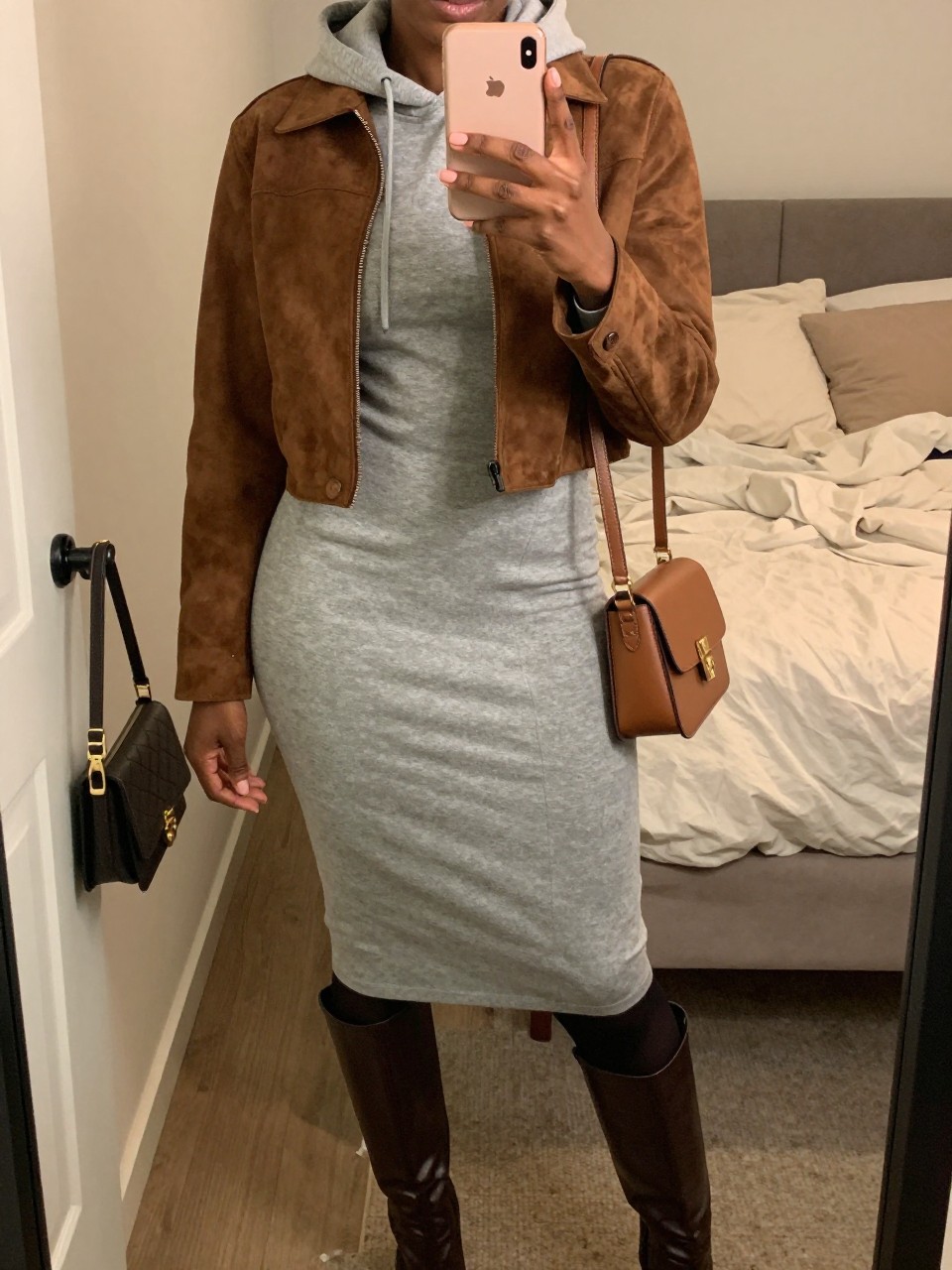 Mirror selfie of a Black woman with rich dark skin wearing a cropped espresso brown suede jacket over a fitted heather gray hoodie midi dress, black opaque tights, dark brown knee-high leather boots, and a compact espresso leather crossbody bag, clearly showing the exact clothing items, colors, textures, and accessories listed, holding a latest-model iPhone in front of a mirror, cropped at the neck so the face does not show, relaxed casual selfie pose, in a feminine-themed bedroom with warm moody lighting, soft pillows on an unmade bed and a handbag hanging on a door handle, realistic natural composition, latest iPhone photo quality.