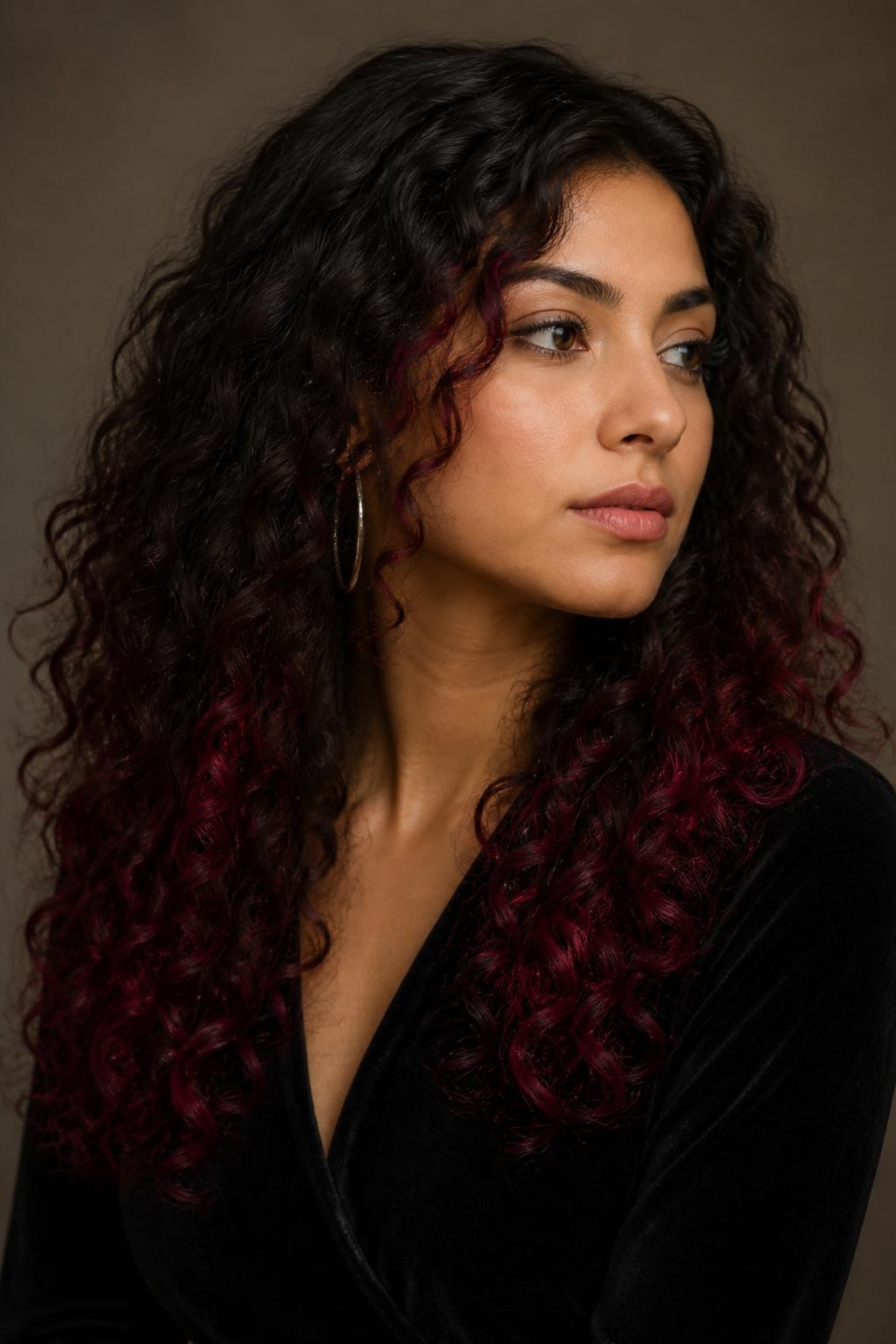 A realistic editorial portrait of a woman with medium olive skin and long corkscrew curls, deep neutral brunette base with garnet-dipped ends, natural dark roots and most mid-lengths preserved, selected curls transitioning through merlot before ending in saturated garnet-red tips, brightest color concentrated around the lower perimeter and two front curls, varying transition heights to avoid a blunt line, no vivid pink or flat burgundy coating, black velvet top, silver hoops, deep taupe background, soft directional light, realistic vivid pigment and natural curl movement.
