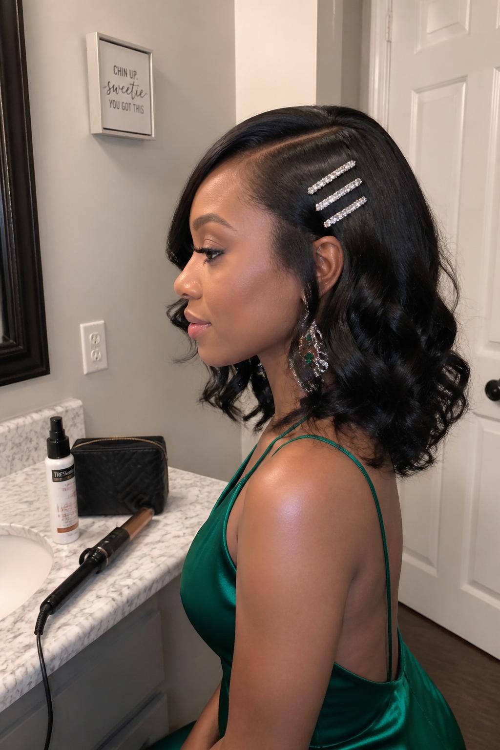 Photo of a Black woman wearing shoulder-length glossy wand curls with a deep side part and three slim crystal pins placed above one ear, side-profile view at a bathroom vanity, warm soft indoor lighting, curling wand cooling beside a heat-protectant bottle and makeup pouch, emerald satin dress visible, statement earring peeking through the curls, realistic shine and natural root texture, latest iPhone photo quality.