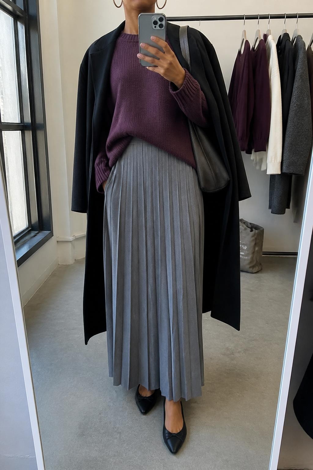 mirror selfie of a Black woman wearing the exact outfit described above, clearly showing a gray pleated maxi skirt, plum oversized knit sweater tucked slightly at the front, black pointed flats, charcoal shoulder bag, silver hoop earrings, and black wool coat draped open, holding a phone in front of a mirror, cropped at NECK so the face does not show, with a relaxed casual selfie pose, in a fashion studio mirror corner, natural window light, with clothing rack with garments behind and a tote bag resting on the floor, latest iPhone photo quality.