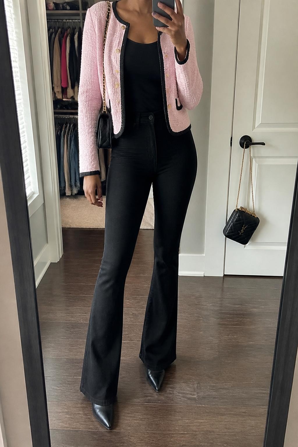 mirror selfie of a Black woman wearing a pale pink bouclé jacket with black trim over a fitted black scoop-neck top, black high-waisted flare jeans, black pointed ankle boots, and a quilted black shoulder bag, clearly showing the same clothing items, colors, fabrics, and accessories listed in the Outfit Pieces section, holding a phone in front of a mirror, cropped at NECK so the face does not show, with a relaxed casual selfie pose, in a walk-in closet mirror, natural window light, including an open closet with clothes visible and a handbag hanging on a door handle, latest iPhone photo quality.