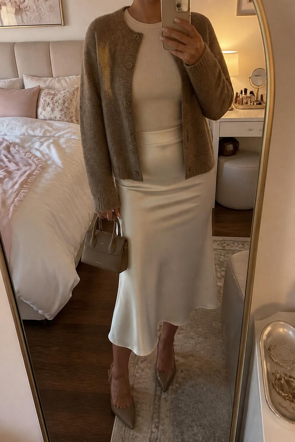 mirror selfie of a woman wearing a mocha cardigan jacket, ivory satin slip skirt, cream fitted knit top, taupe slingback heels, and mocha mini work bag, clearly showing the same clothing items, colors, fabrics, and accessories listed in the Outfit Pieces section, holding a phone in front of a mirror, cropped at NECK so the face does not show, with a relaxed casual selfie pose, in a feminine-themed bedroom, evening golden hour, including soft pillows on an unmade bed and a jewelry tray on the vanity, latest iPhone photo quality.