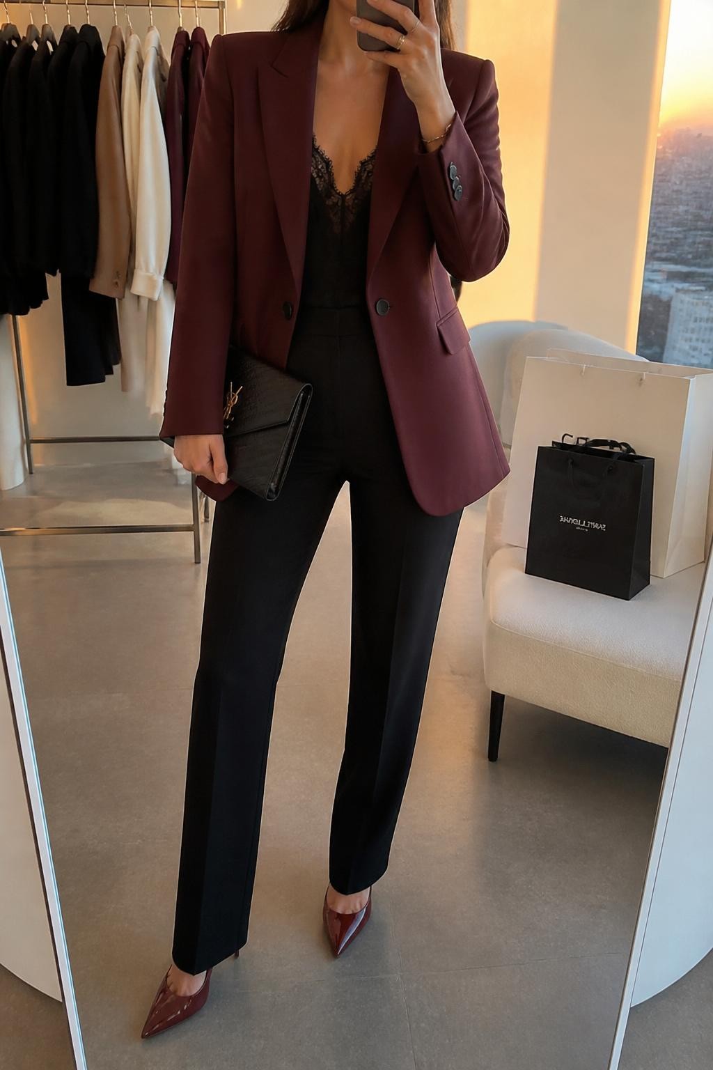 mirror selfie of a woman wearing the exact outfit described above, clearly showing a wine red tailored blazer, black lace top, black straight-leg trousers, red pointed heels, and a black envelope clutch, holding a phone in front of a mirror, cropped at NECK so the face does not show, with a relaxed casual selfie pose, in a fashion studio mirror corner, evening golden hour, with a clothing rack with garments behind and shopping bags beside a chair, latest iPhone photo quality.