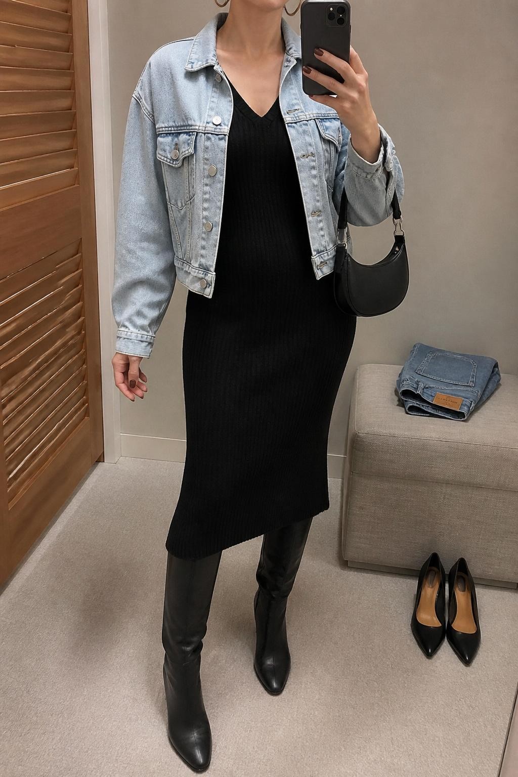 mirror selfie of a woman wearing the exact outfit described above, clearly showing a light-wash denim jacket layered over a black ribbed knit midi dress, black knee-high boots, a black mini shoulder bag, and silver hoop earrings, holding a phone in front of a mirror, cropped at NECK so the face does not show, with a relaxed casual selfie pose, in a minimal dressing room, soft indoor lighting, with folded denim jeans on a bench and a pair of heels on the floor nearby, latest iPhone photo quality.