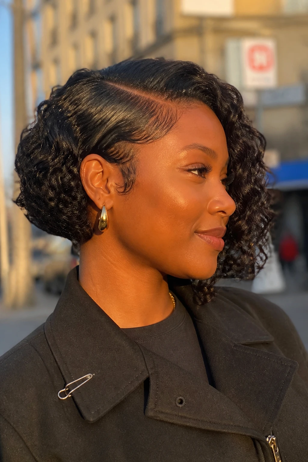 Image prompt

Photo of a Black woman wearing a short curly bob with a deep side part, one side tucked neatly behind the ear, and full defined curls sweeping across the opposite side, side profile view against a softly blurred city street background, golden hour glow, containing a small lived-in detail such as gold hoop earrings and a slim hair clip attached to her jacket collar, realistic candid photograph, latest iPhone photo quality.