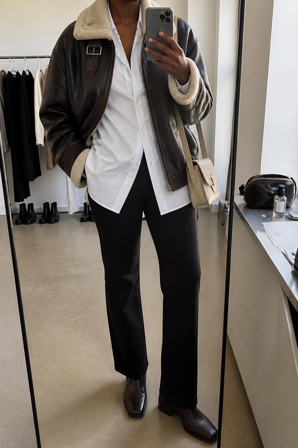 Mirror selfie of a Black woman with rich dark skin wearing an oversized dark brown leather aviator jacket with a cream shearling collar over a long white poplin button-down shirt, black slim straight-leg trousers, dark brown leather ankle boots, and a structured cream shoulder bag, clearly showing the exact clothing items, colors, fabrics, and accessories listed, holding a latest-model iPhone in front of a mirror, cropped at the neck so the face does not show, relaxed casual selfie pose, in a fashion studio mirror corner with natural window light, a clothing rack with garments behind and a cosmetic bag on the counter, natural unstaged composition, latest iPhone photo quality.