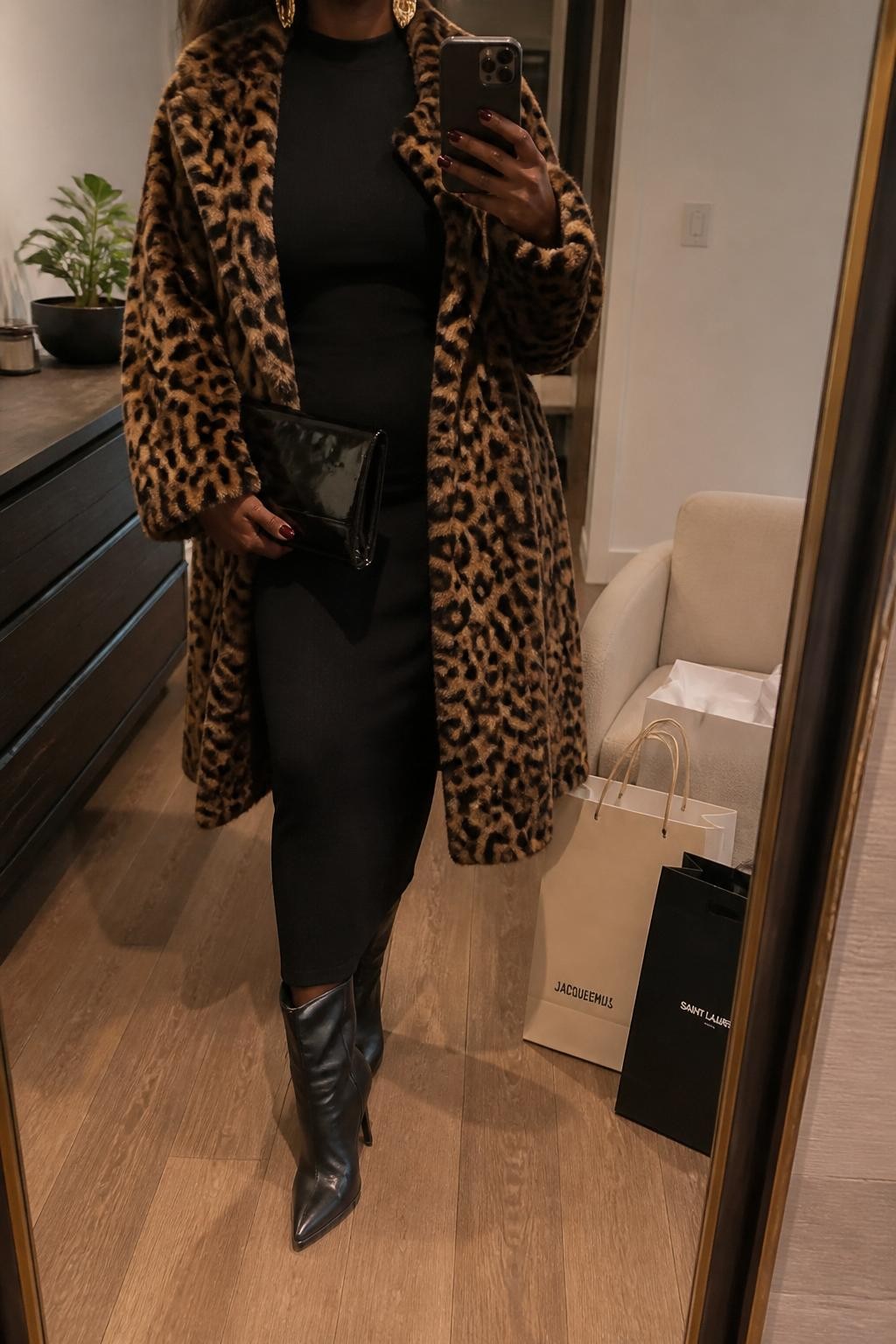 mirror selfie of a Black woman wearing the exact outfit described above, clearly showing a leopard faux fur coat layered over a black fitted midi dress, black heeled boots, a glossy black clutch, and gold statement earrings, holding a phone in front of a mirror, cropped at NECK so the face does not show, with a relaxed casual selfie pose, in a stylish city apartment, warm moody lighting, with shopping bags beside a chair and a small plant on a dresser, latest iPhone photo quality.