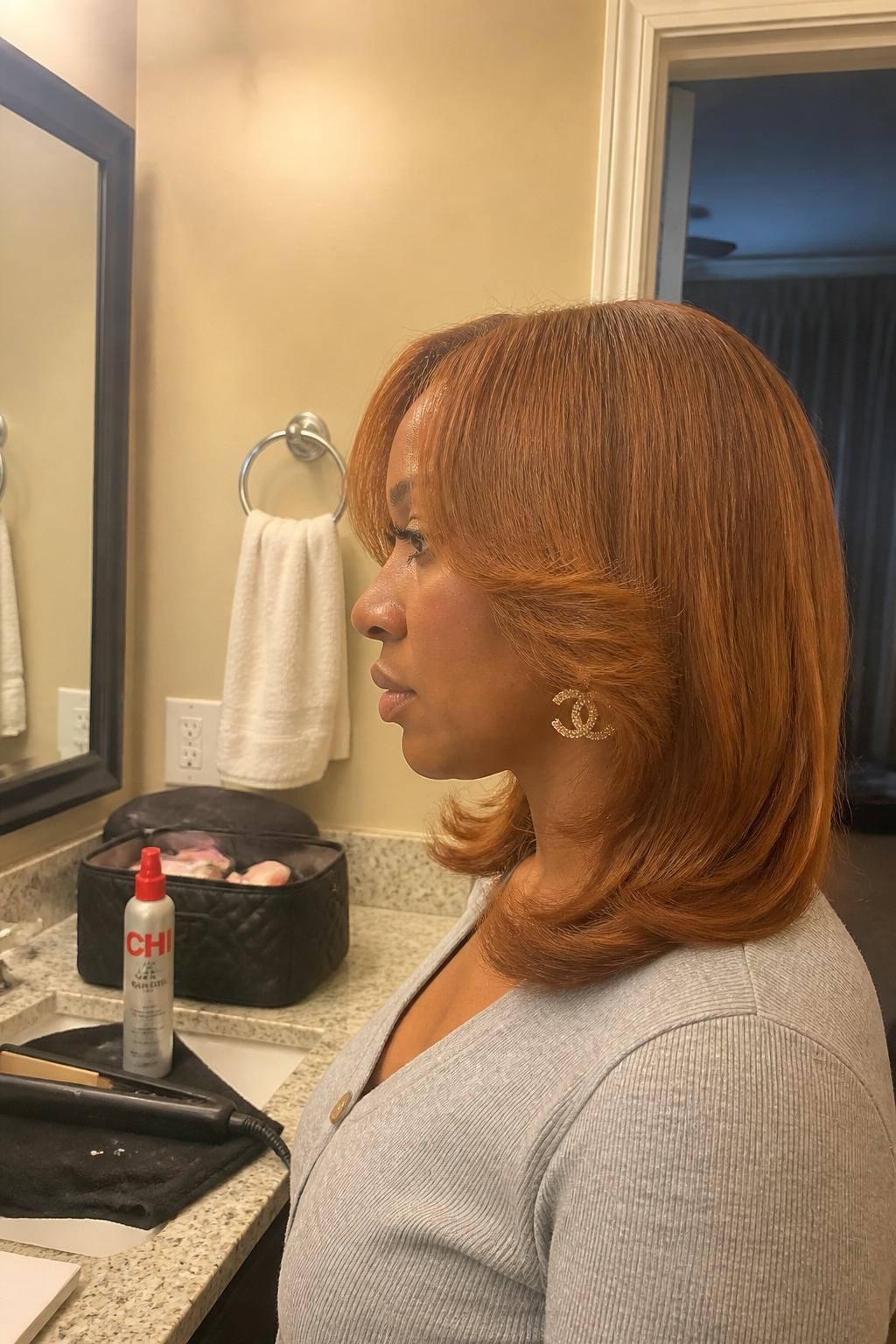 Photo of a Black woman wearing a shoulder-length copper silk press with airy face-framing layers and ends flipped softly away from the face, side-profile view at a bathroom vanity mirror, warm soft indoor lighting, flat iron cooling on the counter beside a heat-protectant bottle and open makeup bag, statement earring peeking through the front layers, natural root texture and realistic shine, latest iPhone photo quality.