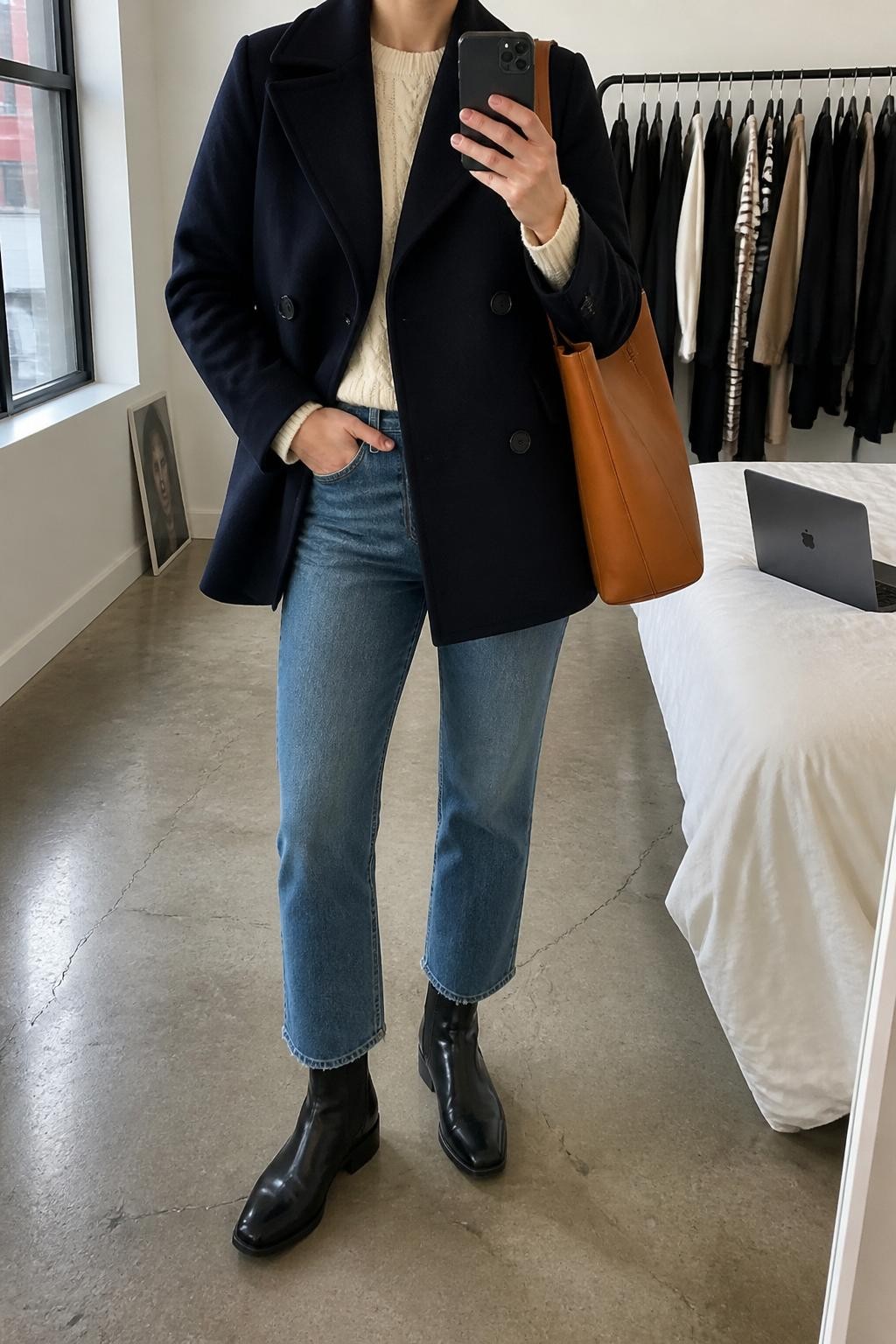 mirror selfie of a woman wearing a navy wool peacoat, cream cable-knit sweater, medium-wash cropped straight jeans, black Chelsea boots, and a tan leather tote bag, clearly showing the same clothing items, colors, fabrics, and accessories listed in the Outfit Pieces section, holding a phone in front of a mirror, cropped at NECK so the face does not show, with a relaxed casual selfie pose, in a fashion studio mirror corner, natural window light, including a clothing rack with garments behind and a laptop on the bed, latest iPhone photo quality.
