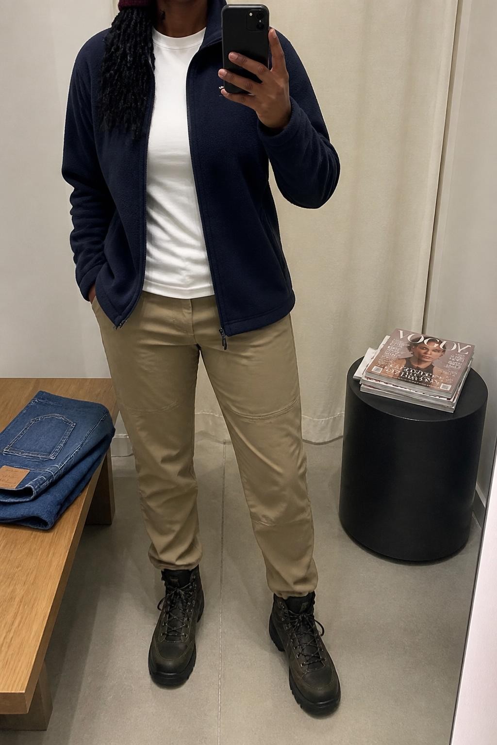 mirror selfie of a Black woman wearing the exact outfit described above, clearly showing a navy fleece zip jacket, white moisture-wicking tee, khaki hiking pants, black waterproof hiking boots, and a burgundy knit beanie, holding a phone in front of a mirror, cropped at NECK so the face does not show, with a relaxed casual selfie pose, in a minimal dressing room, soft indoor lighting, with folded denim jeans on a bench and a stack of fashion magazines on a side table, latest iPhone photo quality.