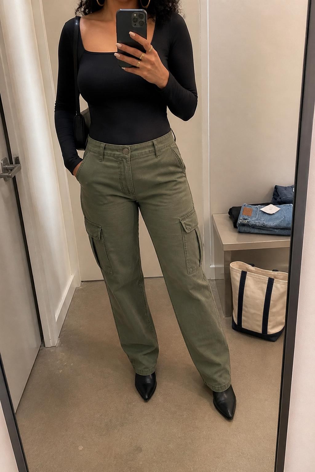 mirror selfie of an Afro-Latina woman wearing olive green cargo pants, a fitted black long-sleeve bodysuit, black pointed ankle boots, chunky gold hoops, and a black shoulder bag, holding a phone in front of a mirror, cropped at NECK so the face does not show, relaxed casual selfie pose, in a minimal dressing room with soft indoor lighting, including folded denim jeans on a bench and a tote bag resting on the floor, realistic latest iPhone photo quality.