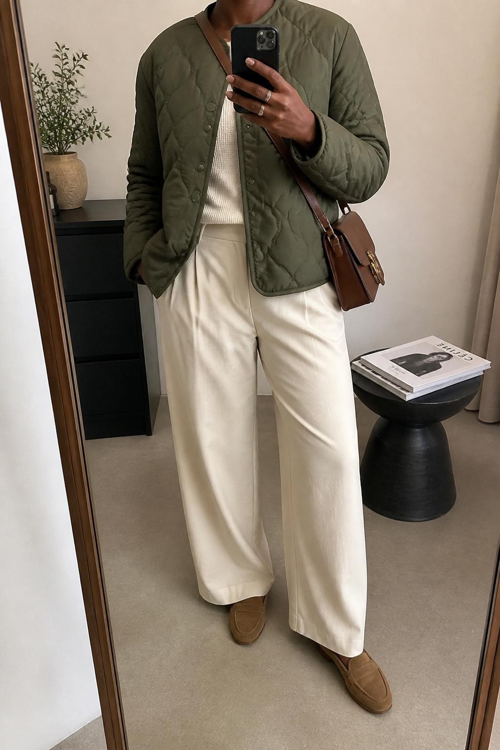 mirror selfie of a Black woman wearing an olive quilted jacket, cream lightweight knit top, ecru wide-leg trousers, tan suede loafers, and a brown leather crossbody bag, clearly showing the same clothing items, colors, fabrics, and accessories listed in the Outfit Pieces section, holding a phone in front of a mirror, cropped at NECK so the face does not show, with a relaxed casual selfie pose, in a minimal dressing room, soft indoor lighting, including a small plant on a dresser and a stack of fashion magazines on a side table, latest iPhone photo quality.