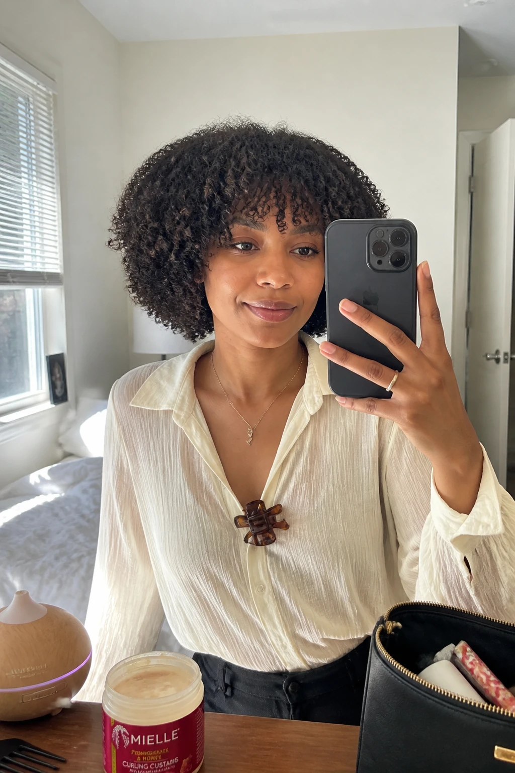 Image prompt

Photo of a woman wearing a chin-length 4A curly bob with dense defined coils, full soft bangs resting above the eyebrows, and a rounded shape framing the jaw, front-facing candid mirror selfie in a bright bedroom, natural window light, containing a small lived-in detail such as a diffuser and curl cream jar on the dresser, a makeup bag nearby, and a claw clip clipped to her blouse collar, realistic everyday getting-ready moment, latest iPhone photo quality.