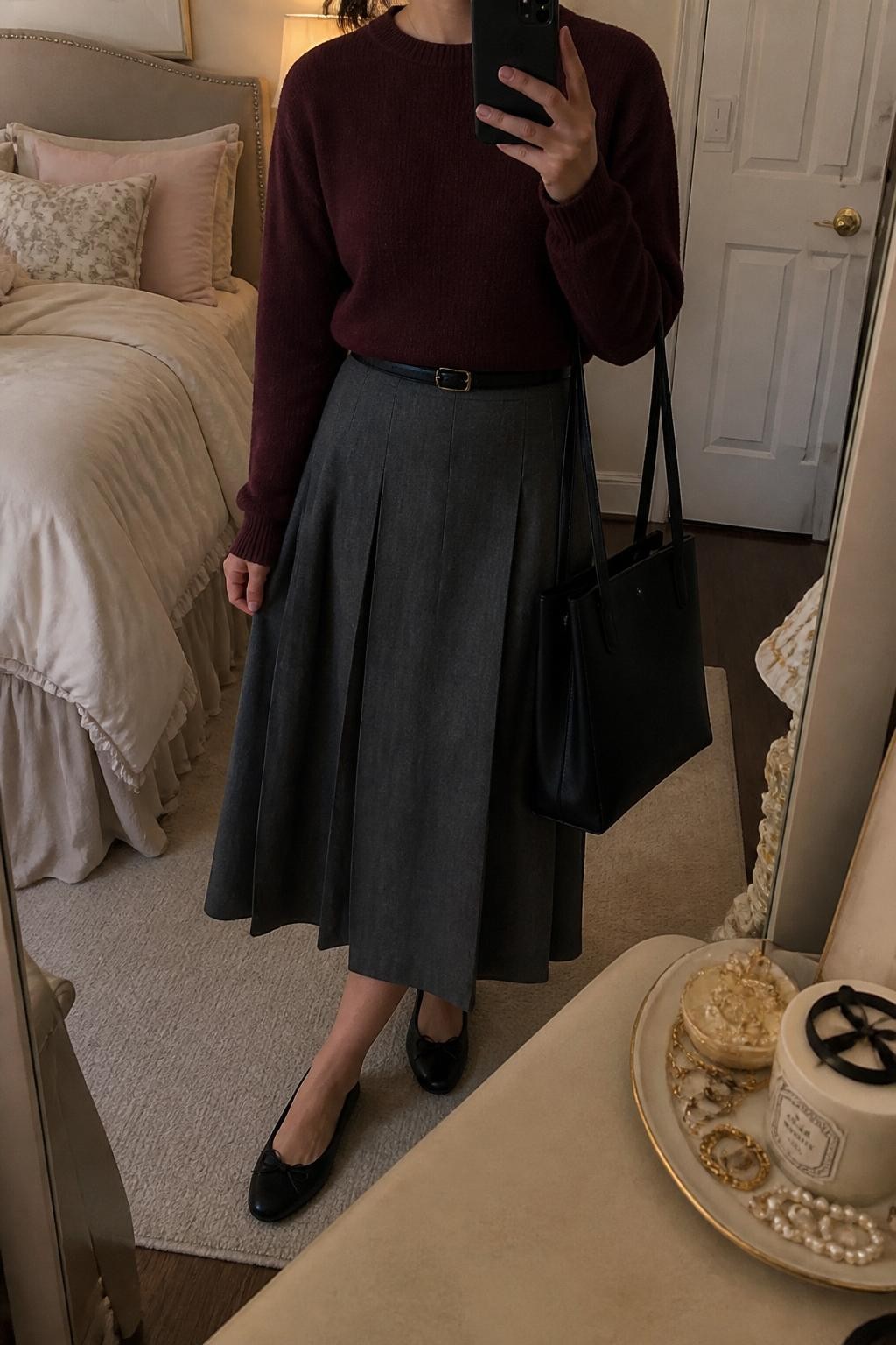 mirror selfie of a woman wearing a burgundy soft knit sweater tucked into a gray pleated midi skirt, black ballet flats, slim black belt, and a structured black tote bag, clearly showing the same clothing items, colors, fabrics, and accessories listed in the Outfit Pieces section, holding a phone in front of a mirror, cropped at NECK so the face does not show, with a relaxed casual selfie pose, in a feminine-themed bedroom, warm moody lighting, including a jewelry tray on the vanity and soft pillows on an unmade bed, latest iPhone photo quality.