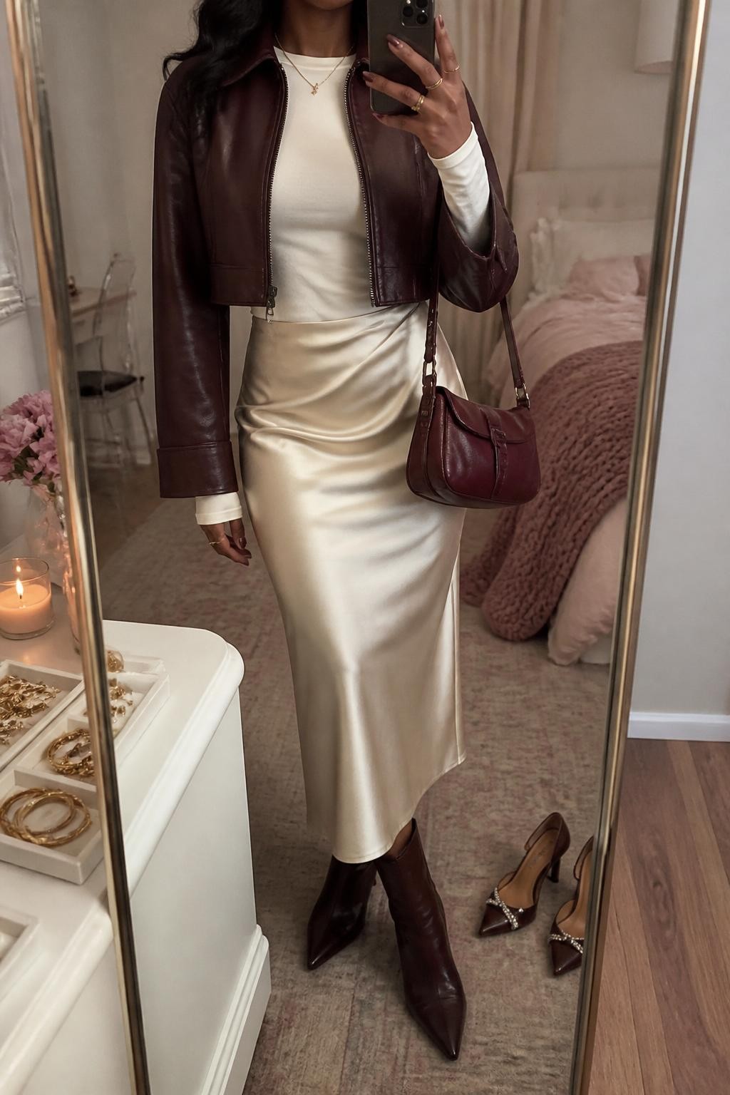 mirror selfie of a Black woman wearing a cropped burgundy leather jacket, fitted cream long-sleeve top, champagne cream satin midi skirt, chocolate brown pointed ankle boots, and a small burgundy shoulder bag, clearly showing the same clothing items, colors, fabrics, and accessories listed in the Outfit Pieces section, holding a phone in front of a mirror, cropped at NECK so the face does not show, with a relaxed casual selfie pose, in a feminine-themed bedroom, warm moody lighting, including a jewelry tray on the vanity and a pair of heels on the floor nearby, latest iPhone photo quality.