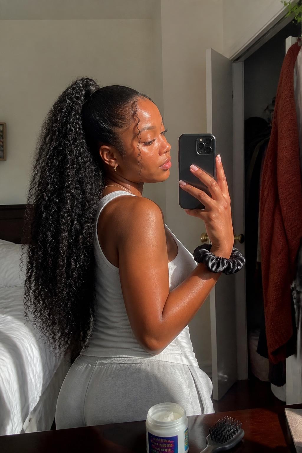 Photo of a Black woman wearing very long natural curls styled into a high ponytail with full defined texture, softly shaped edges, and two curled tendrils near the temples, candid side-profile mirror selfie in a sun-drenched bedroom, natural window light, curl cream and edge brush resting on the dresser, silk scrunchie around her wrist and a rust-colored cardigan hanging inside a slightly open closet, realistic getting-ready moment, latest iPhone photo quality.