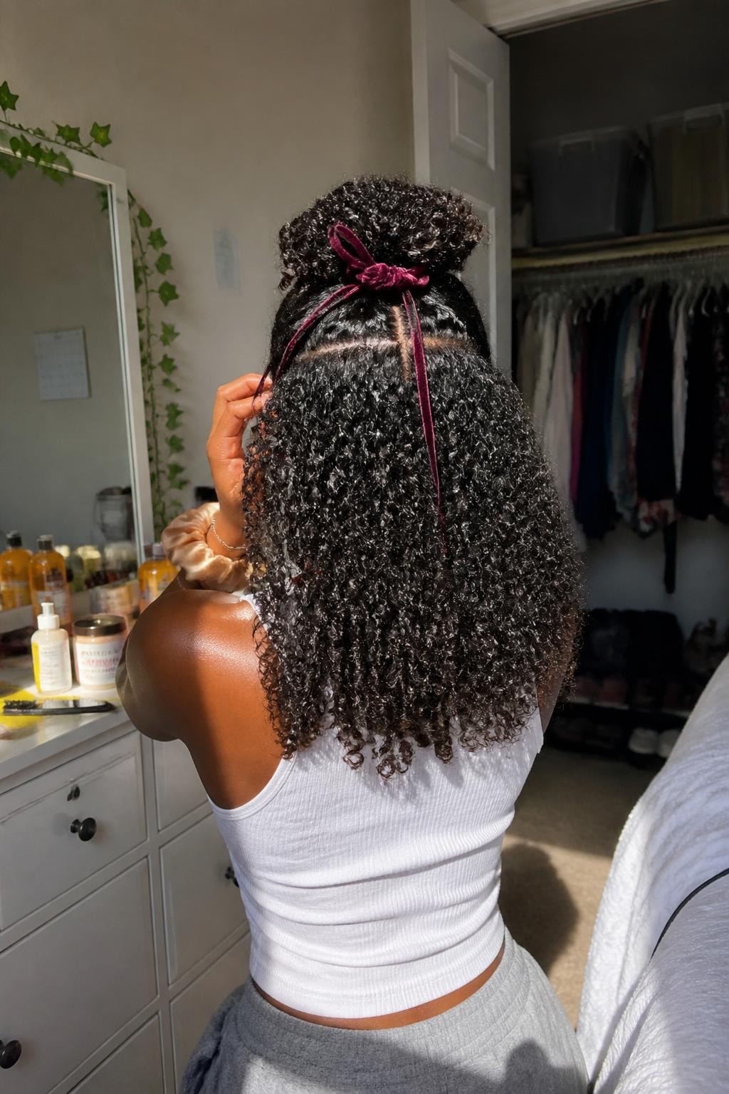 Photo of a Black woman wearing medium-length natural curls styled into a half-up ponytail with two slim front braids leading toward the crown and a burgundy velvet ribbon wrapped around the base, back-of-head view in a sun-drenched bedroom, warm natural window light, edge brush and curl cream on the dresser, silk scrunchie around her wrist and an open closet behind her, realistic getting-ready moment, latest iPhone photo quality.