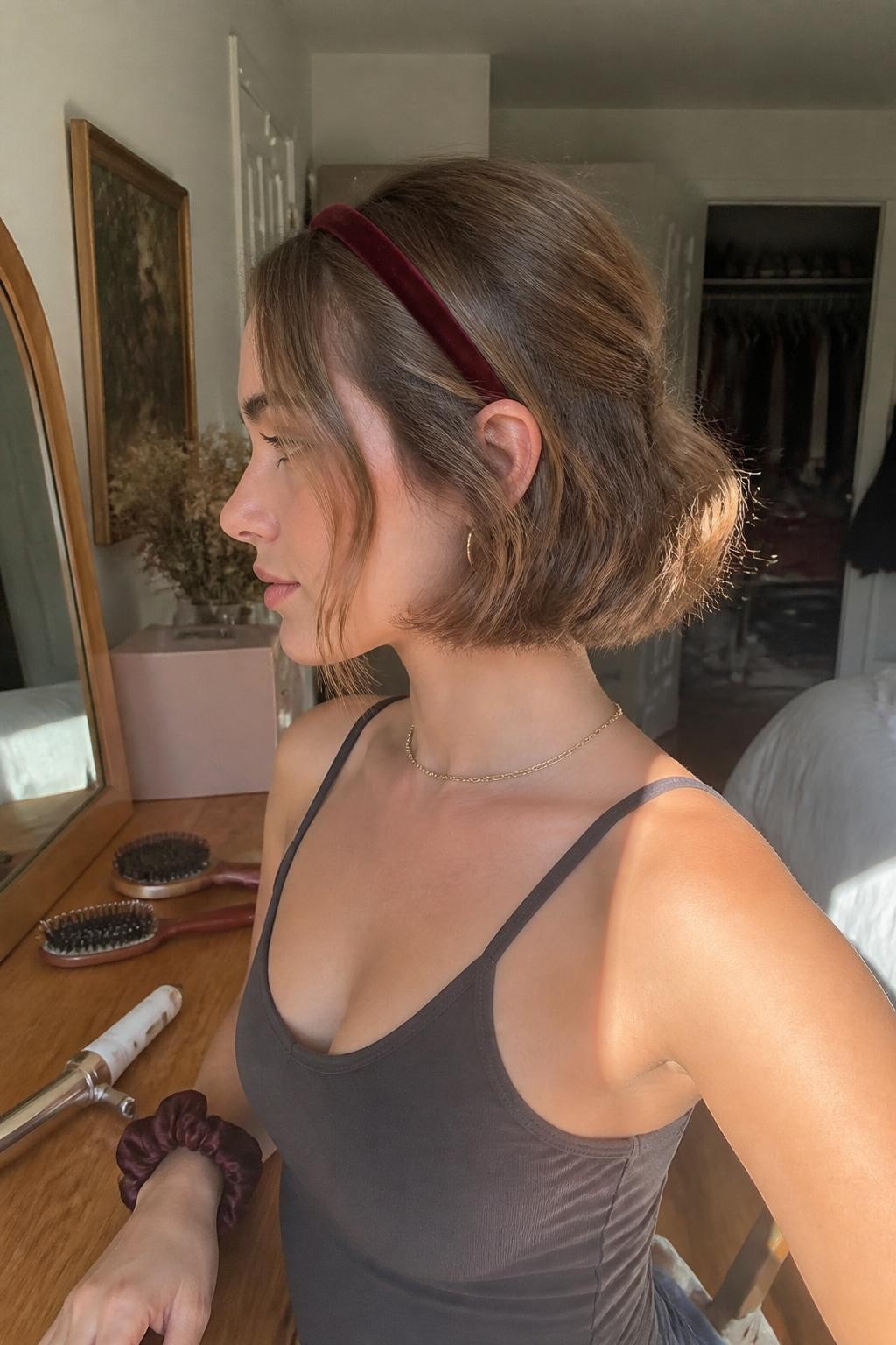 Photo of a woman wearing a jaw-length wavy bob with a narrow burgundy velvet headband placed behind the hairline, softly bent ends, and loose pieces around the ears, side-profile view in a sun-drenched bedroom, warm natural window light, curling wand and hairbrush on the dresser, a silk scrunchie around her wrist and a slightly open closet behind her, latest iPhone photo quality.