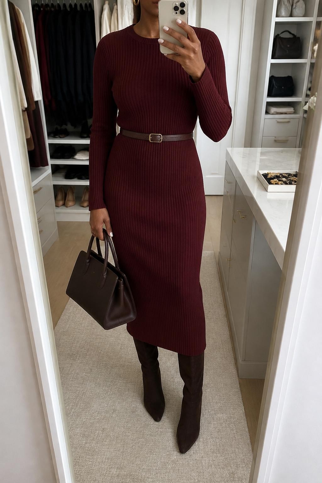 Mirror selfie of a Black woman with deep brown skin wearing a long-sleeved burgundy ribbed-knit midi dress, knee-high chocolate brown suede boots, a slim dark brown leather belt around the waist, small gold hoop earrings, and a structured chocolate leather handbag, clearly showing the exact clothing items, colors, textures, and accessories listed, holding a latest-model iPhone in front of a mirror, cropped at the neck so the face does not show, relaxed casual selfie pose, in a walk-in closet mirror with soft indoor lighting, an open closet with clothes visible and a jewelry tray on the vanity, natural uncluttered setting, latest iPhone photo quality.