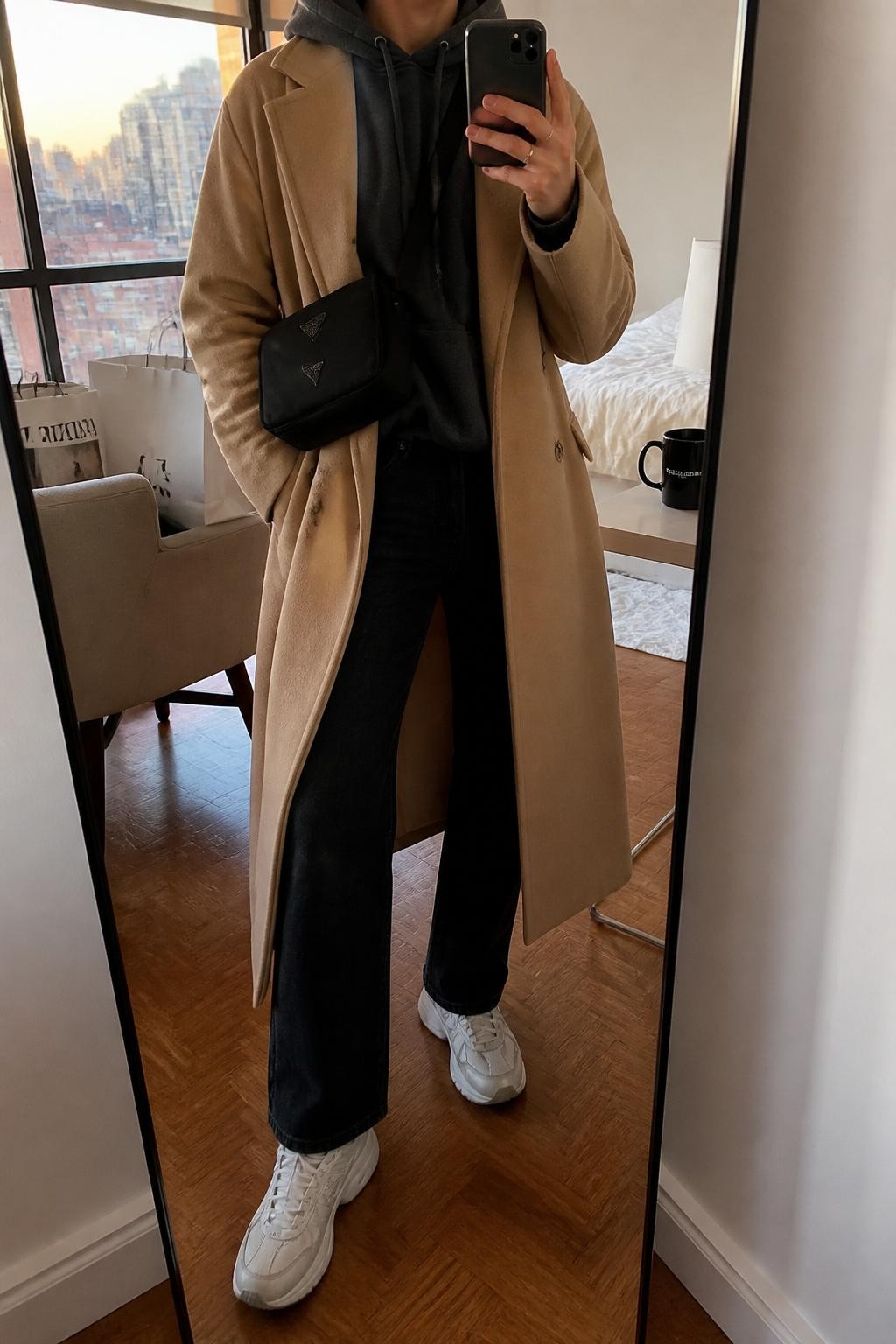 mirror selfie of a woman wearing the exact outfit described above, clearly showing a camel long coat, charcoal hoodie, black straight-leg jeans, white chunky sneakers, and a black nylon crossbody bag, holding a phone in front of a mirror, cropped at NECK so the face does not show, with a relaxed casual selfie pose, in a stylish city apartment, evening golden hour, with shopping bags beside a chair and a coffee mug on a bedside table, latest iPhone photo quality.