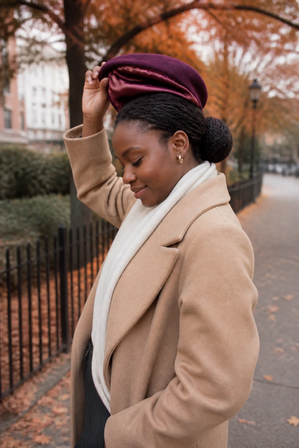 Image Prompt: A realistic outdoor fall fashion and haircare photograph of a Black woman with rich dark skin wearing a satin-lined burgundy beret over a loose low braided bun, natural hairline left relaxed without heavy edge control, woman gently lifting the beret to show the smooth satin lining, camel wool coat, cream scarf, dark trousers, and small gold hoop earrings, copper leaves along a quiet city path, soft overcast daylight, realistic coily hair texture, skin, wool, and satin details, polished DSLR lifestyle photography, 70mm lens, no fashion logos, no text.