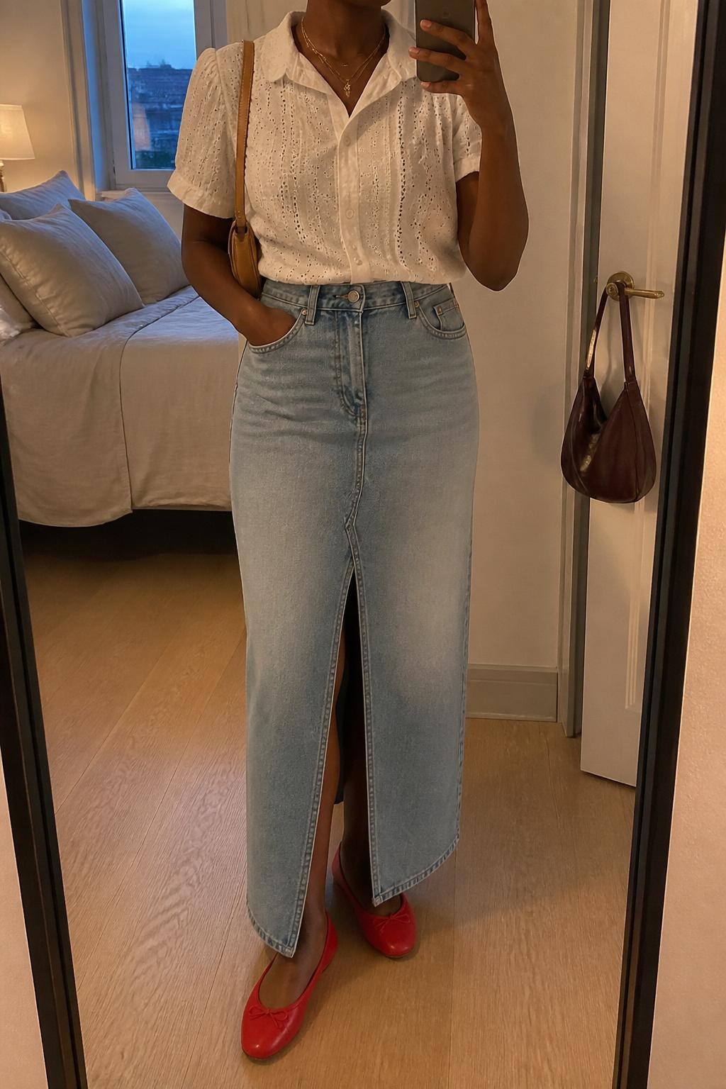 mirror selfie of a Black woman wearing a white eyelet short-sleeve blouse tucked into a light wash denim maxi skirt with front slit, red ballet flats, tan mini shoulder bag, and gold layered necklaces, clearly showing the same clothing items, colors, fabrics, and accessories listed in the Outfit Pieces section, holding a phone in front of a mirror, cropped at NECK so the face does not show, with a relaxed casual selfie pose, in a cozy bedroom mirror setup, evening golden hour, including soft pillows on an unmade bed and a handbag hanging on a door handle, latest iPhone photo quality.