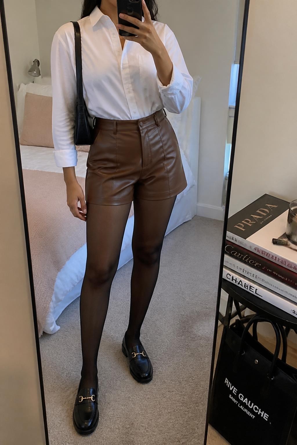 mirror selfie of a Latina woman wearing a crisp white button-down shirt, brown faux leather tailored shorts, sheer black tights, black loafers, and a black shoulder bag, holding a phone in front of a mirror, cropped at NECK so the face does not show, relaxed casual selfie pose, in a cozy bedroom mirror setup with soft indoor lighting, including a tote bag resting on the floor and a stack of fashion magazines on a side table, realistic latest iPhone photo quality.