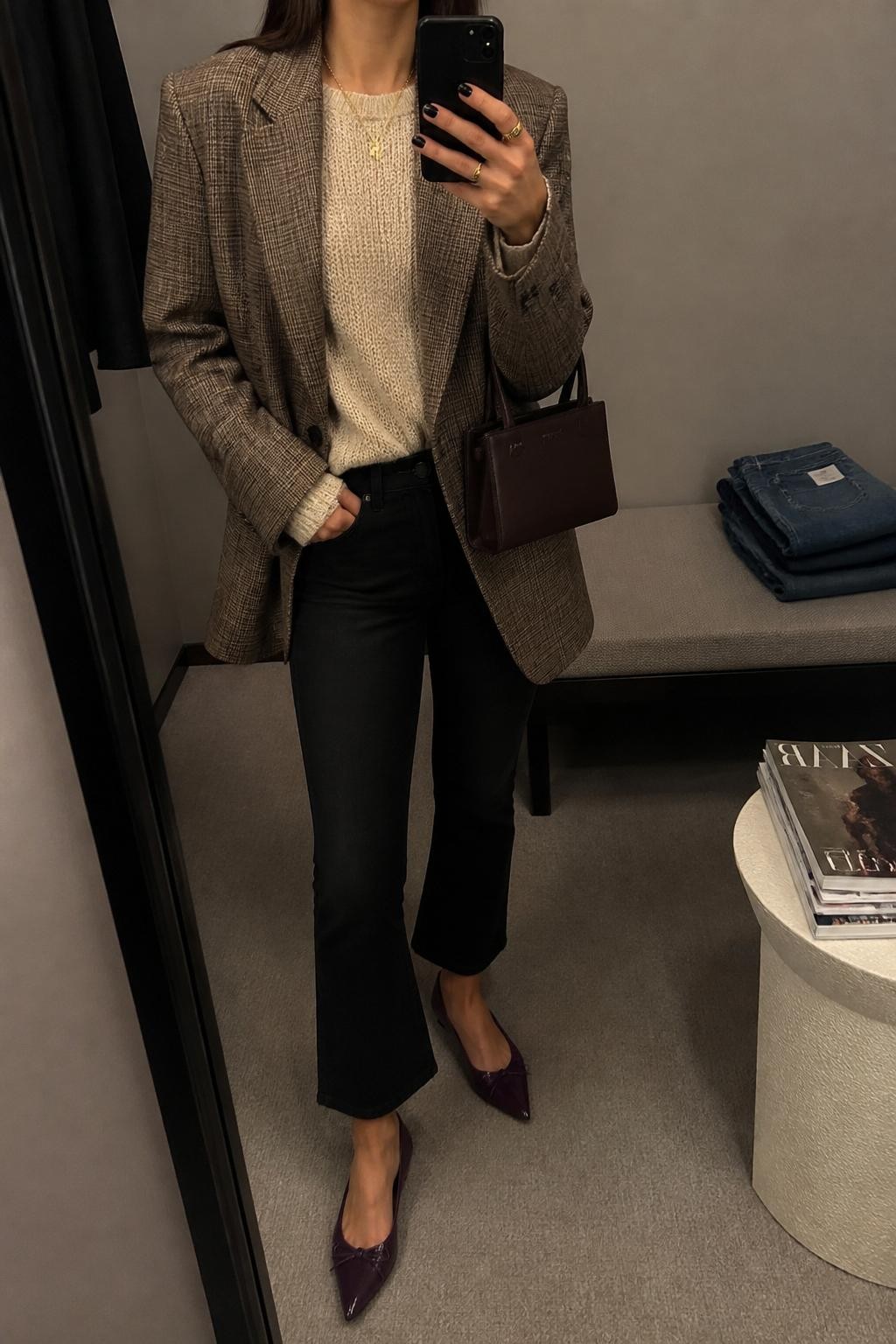 mirror selfie of a woman wearing the exact outfit described above, clearly showing a brown plaid oversized blazer layered over a cream fine-knit sweater, black cropped flare jeans, violet pointed flats, a chocolate brown mini handbag, and gold pendant necklace, holding a phone in front of a mirror, cropped at NECK so the face does not show, with a relaxed casual selfie pose, in a minimal dressing room, warm moody lighting, with folded denim jeans on a bench and a stack of fashion magazines on a side table, latest iPhone photo quality.