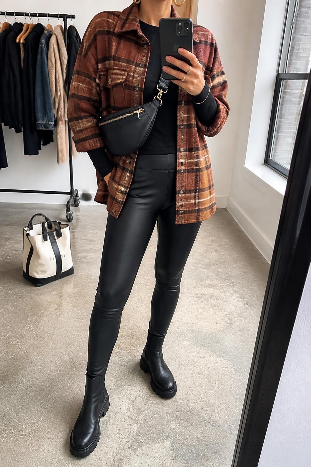 mirror selfie of a woman wearing the exact outfit described above, clearly showing a rust and brown plaid wool shacket layered over a fitted black long-sleeve top, black leather leggings, black lug-sole boots, a black belt bag worn crossbody, and gold hoop earrings, holding a phone in front of a mirror, cropped at NECK so the face does not show, with a relaxed casual selfie pose, in a fashion studio mirror corner, natural window light, with clothing rack with garments behind and a tote bag resting on the floor, latest iPhone photo quality.