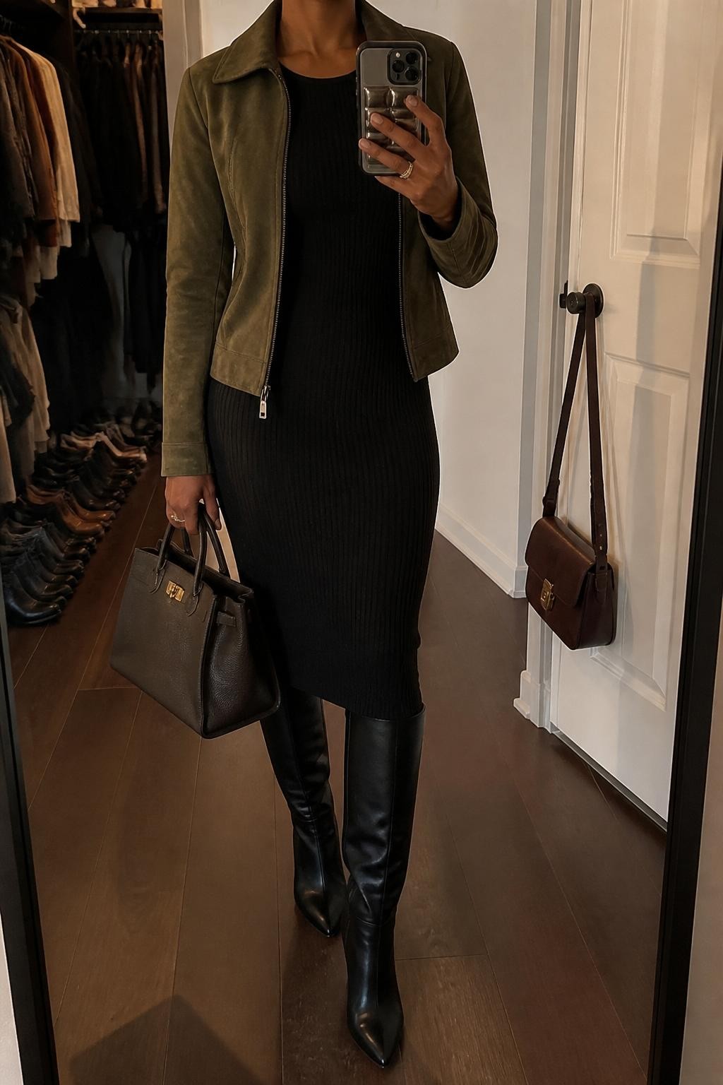 mirror selfie of a Black woman wearing an olive green suede jacket, fitted black ribbed midi dress, black leather knee-high boots, small gold hoop earrings, and a structured dark brown handbag, clearly showing the same clothing items, colors, fabrics, and accessories listed in the Outfit Pieces section, holding a phone in front of a mirror, cropped at NECK so the face does not show, with a relaxed casual selfie pose, in a walk-in closet mirror, evening golden hour, including an open closet with clothes visible and a handbag hanging on a door handle, latest iPhone photo quality.