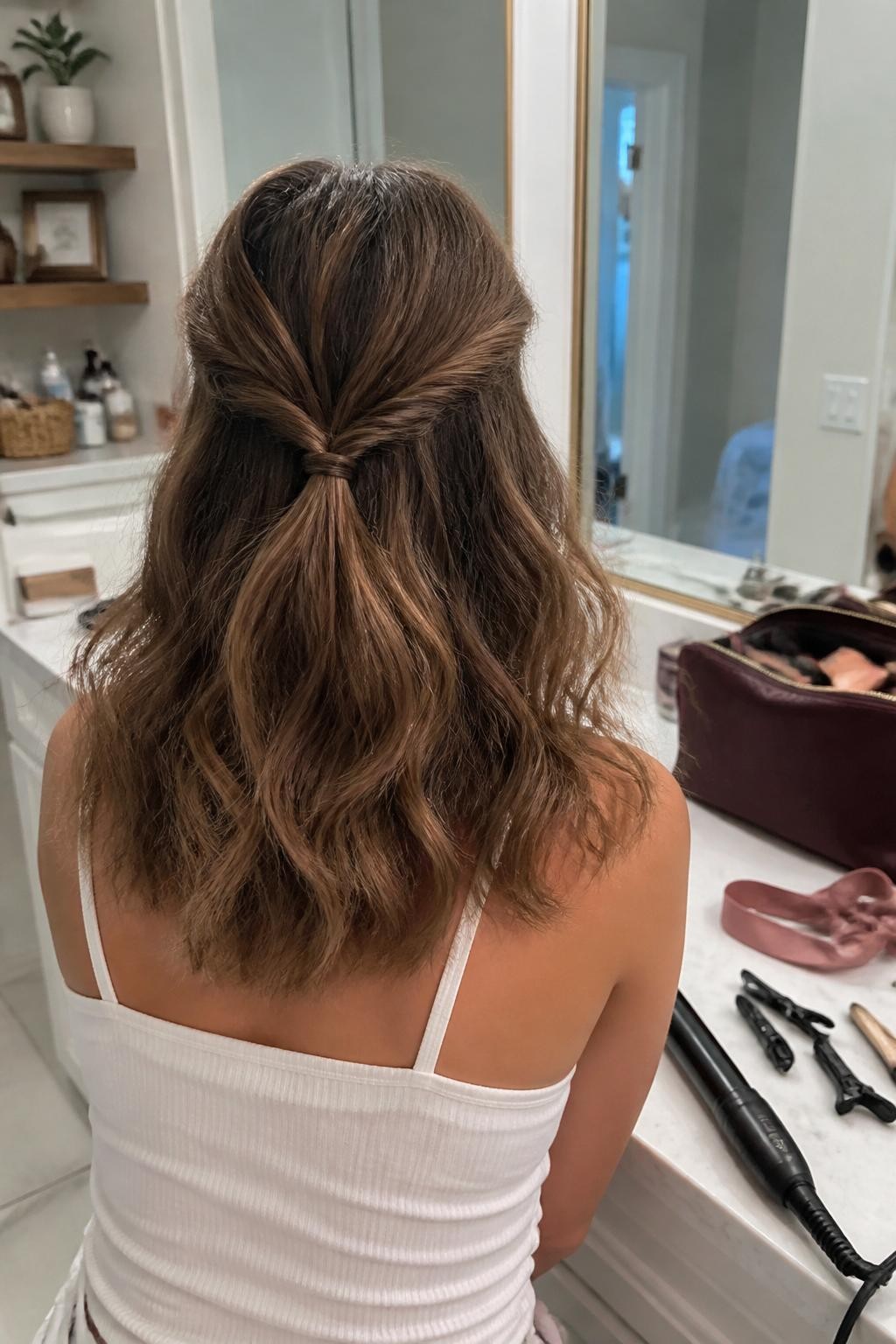 Image prompt: Photo of a woman wearing loose shoulder-length waves with two small rope braids meeting in a half-up ponytail, back-of-head view clearly showing the twisted texture, bathroom vanity mirror setting, soft indoor lighting, curling wand and several sectioning clips on the counter, a velvet ribbon beside the makeup bag, natural flyaways, latest iPhone photo quality.