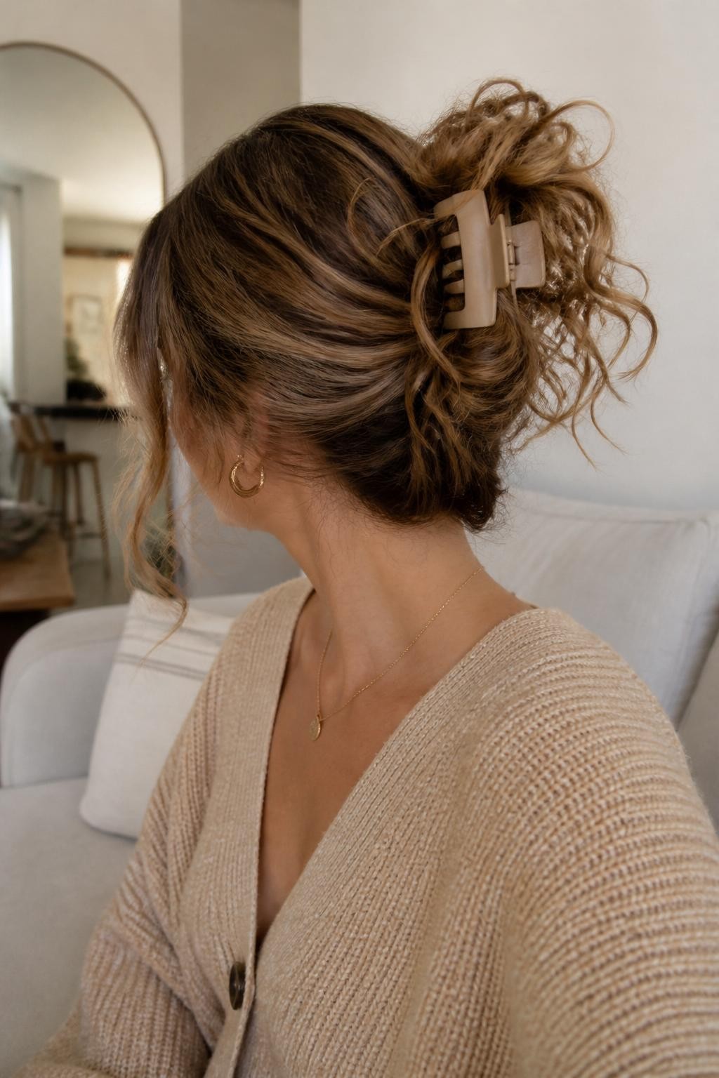 A realistic editorial hairstyle photo of a woman with long honey brown curly hair styled into a messy claw clip bun, curls loosely gathered at the back with a matte beige claw clip, curly ends spilling over the clip, soft front pieces left out, oversized cardigan, natural morning light, relaxed apartment background, casual cozy hair inspiration, high-resolution DSLR photo, no text, (no watermarks on images)