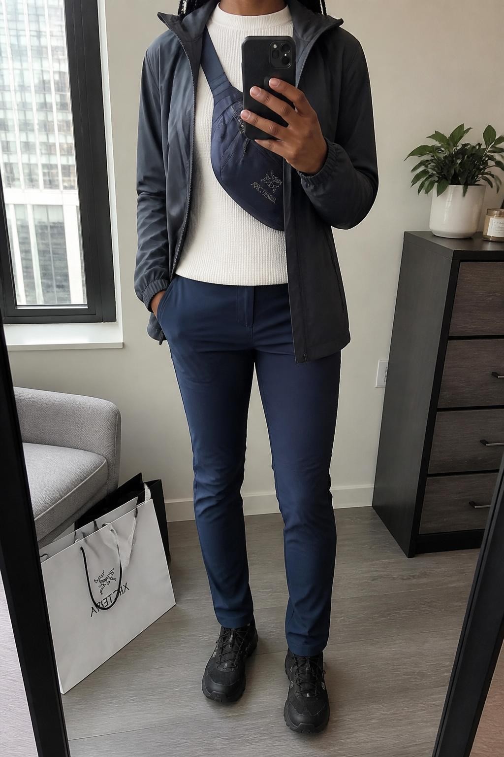 mirror selfie of a Black woman wearing the exact outfit described above, clearly showing a charcoal softshell hiking jacket, navy slim trail pants, white thermal crewneck top, black trail sneakers, and a navy crossbody hiking sling bag, holding a phone in front of a mirror, cropped at NECK so the face does not show, with a relaxed casual selfie pose, in a stylish city apartment, natural window light, with shopping bags beside a chair and a small plant on a dresser, latest iPhone photo quality.