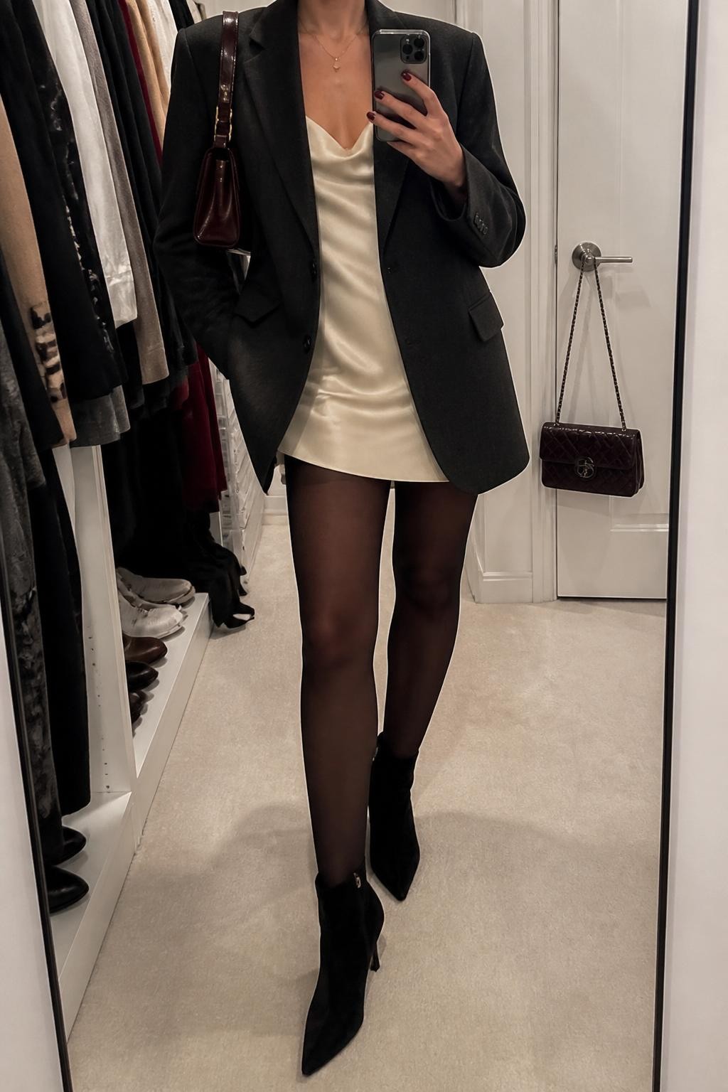 mirror selfie of a woman wearing an oversized charcoal wool blazer, ivory satin slip dress, sheer black tights, black suede ankle boots, and a small burgundy leather bag, clearly showing the same clothing items, colors, fabrics, and accessories listed in the Outfit Pieces section, holding a phone in front of a mirror, cropped at NECK so the face does not show, with a relaxed casual selfie pose, in a walk-in closet mirror, soft indoor lighting, including an open closet with clothes visible and a handbag hanging on a door handle, latest iPhone photo quality.