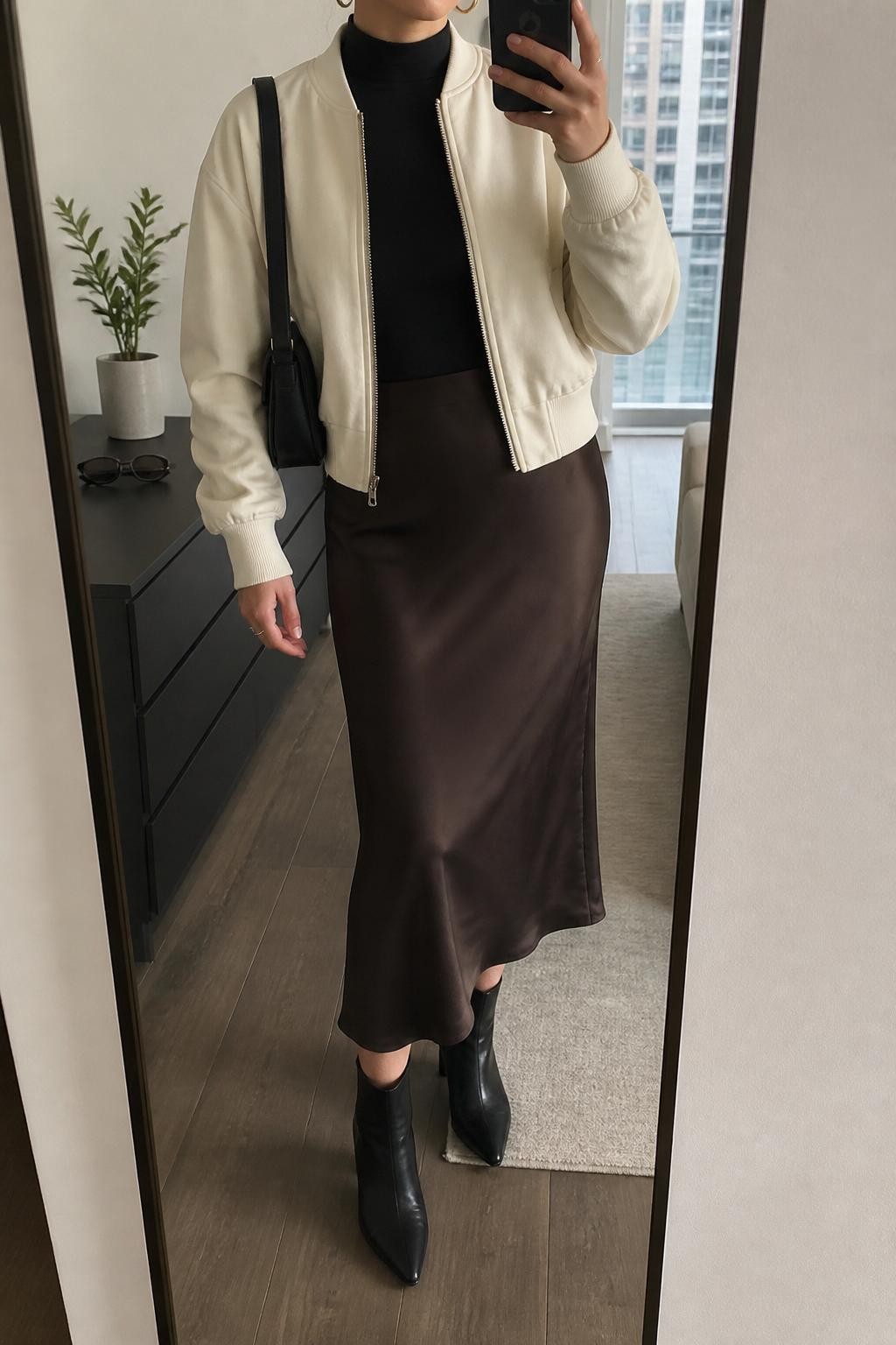 mirror selfie of a woman wearing the exact outfit described above, clearly showing an ivory bomber jacket layered over a fitted black mock-neck top, dark espresso satin midi skirt, black heeled ankle boots, a black leather shoulder bag, and gold huggie earrings, holding a phone in front of a mirror, cropped at NECK so the face does not show, with a relaxed casual selfie pose, in a stylish city apartment, natural window light, with a small plant on a dresser and sunglasses placed on a dresser, latest iPhone photo quality.