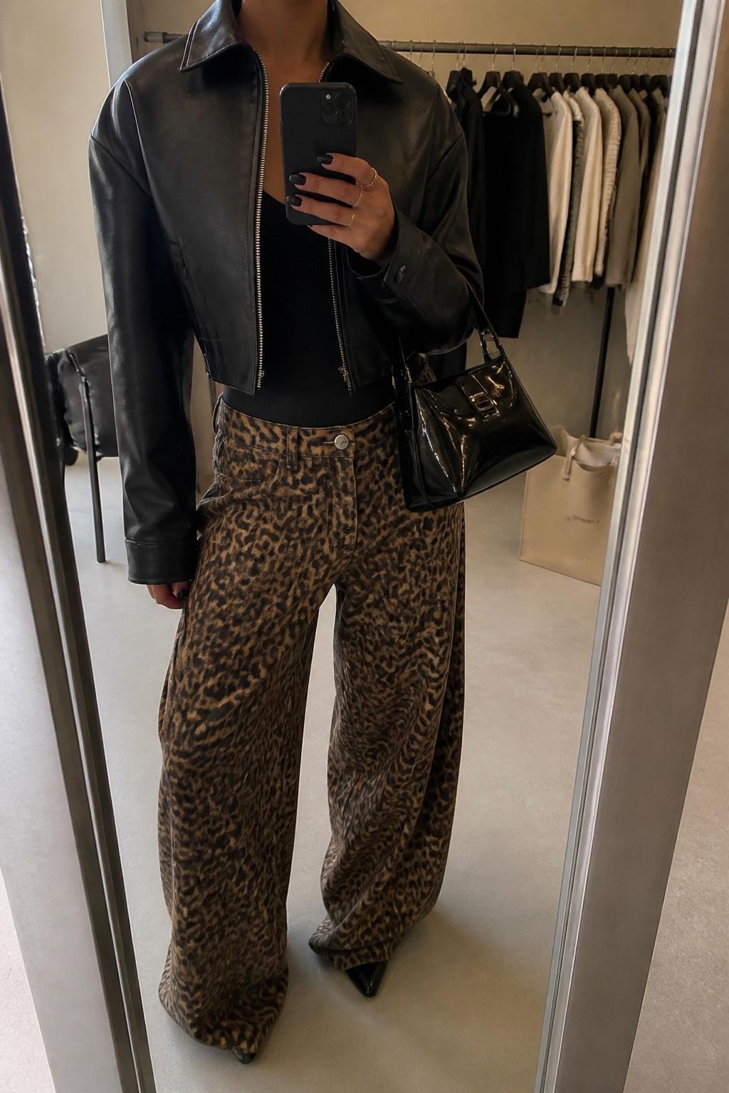 mirror selfie of a woman wearing the exact outfit described above, clearly showing a cropped black leather jacket, fitted black bodysuit, leopard wide-leg pants, black pointed heels, and a glossy black shoulder bag, holding a phone in front of a mirror, cropped at NECK so the face does not show, with a relaxed casual selfie pose, in a fashion studio mirror corner, warm moody lighting, with a clothing rack with garments behind and a tote bag resting on the floor, latest iPhone photo quality.