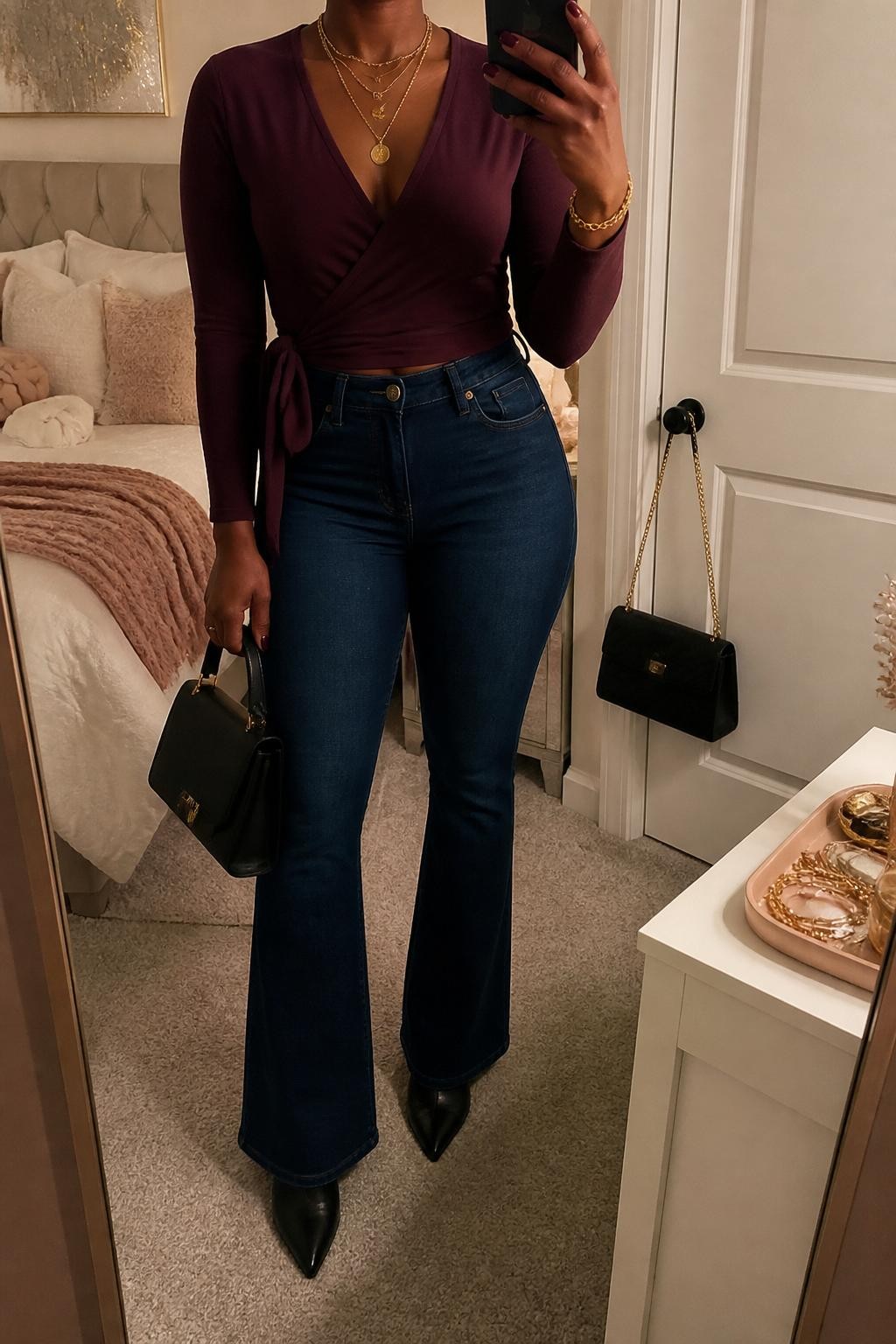 mirror selfie of a Black woman wearing the exact outfit described above, clearly showing a plum wrap top with long sleeves, dark blue flare jeans, black pointed ankle boots, a black structured handbag, and gold layered necklaces, holding a phone in front of a mirror, cropped at NECK so the face does not show, with a relaxed casual selfie pose, in a feminine-themed bedroom, evening golden hour, with a handbag hanging on a door handle and a jewelry tray on the vanity, latest iPhone photo quality.