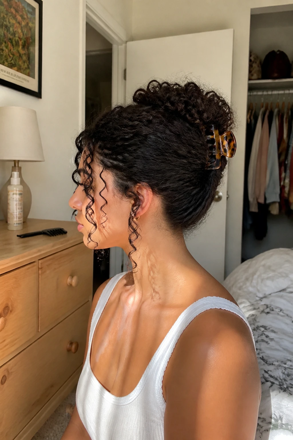 Image prompt
Photo of a woman wearing long 3B curls in a claw clip twist with the hair folded upward, soft ringlets spilling over the top of a large tortoiseshell clip, and two loose curls around the face, side profile view in a sun-drenched bedroom, natural window light, containing a small lived-in detail such as a wide-tooth comb on the dresser, a curl spray bottle nearby, and a slightly open closet behind her, realistic getting-ready moment, latest iPhone photo quality.
