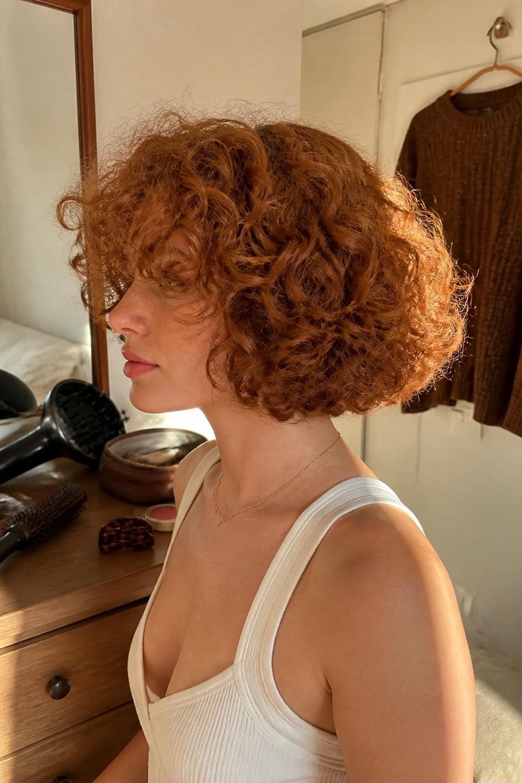Photo of a woman wearing a chin-length copper curly bob with rounded volume, fluffy curly bangs grazing the eyebrows, and softly defined ends, side-profile view in a sun-drenched bedroom, warm window light, diffuser and curl brush on the dresser, tortoiseshell clip beside an open makeup bag and a brown cardigan hanging nearby, realistic mixed curl pattern and natural frizz, latest iPhone photo quality.