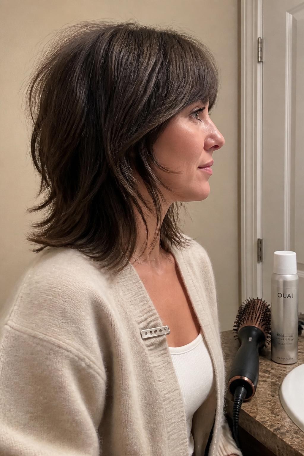Photo of a woman wearing a shoulder-length modern shag with choppy feathered layers, bottleneck bangs, and slightly flicked ends, side-profile view at a bathroom vanity, soft indoor lighting, blow-dry brush and texture spray on the counter, a small metal hair clip clipped to the collar of her cardigan, natural separation between the layers, latest iPhone photo quality.